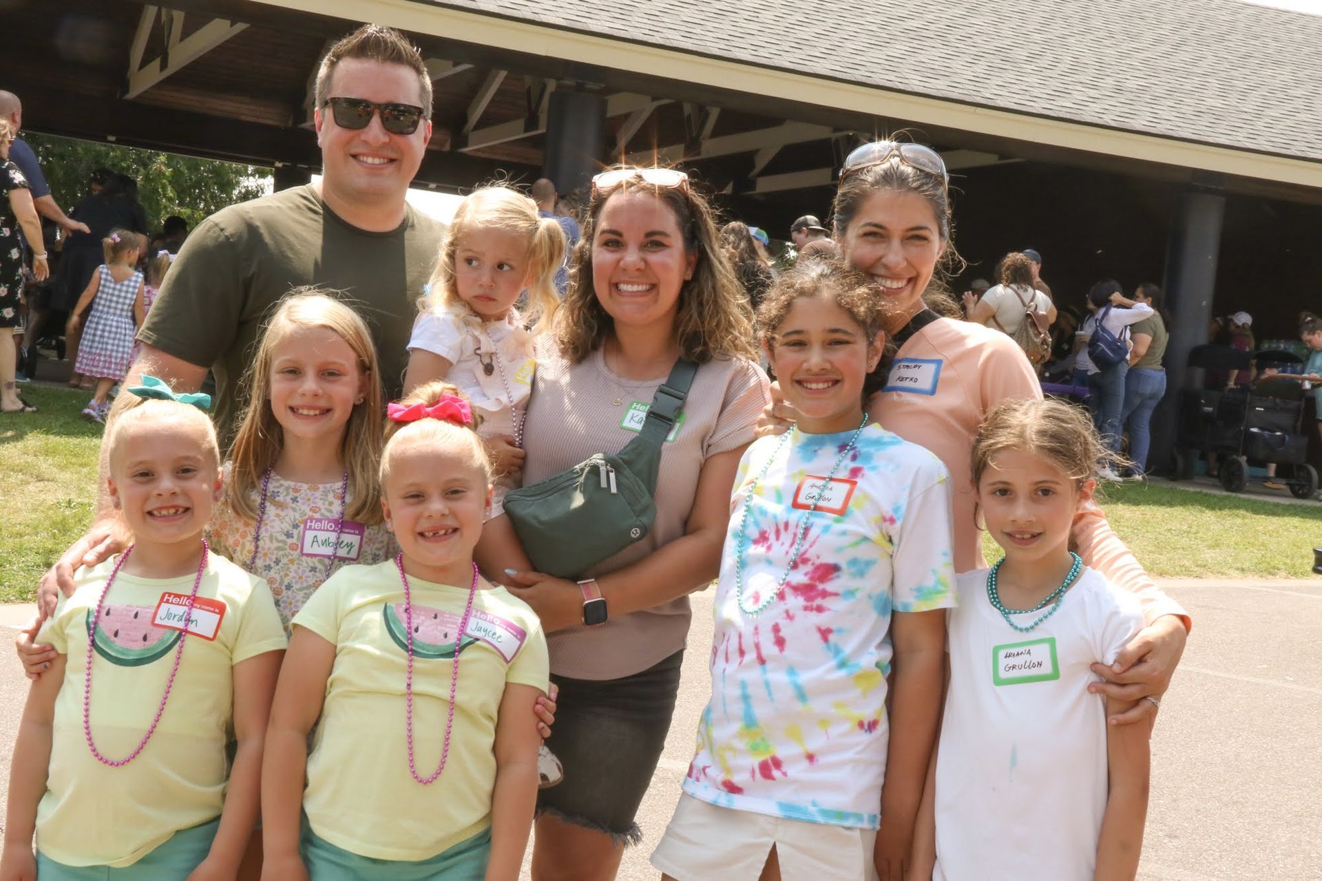 Group of people smiling for a photo outdoors. Some children wear watermelon necklaces and tie-dye shirts.