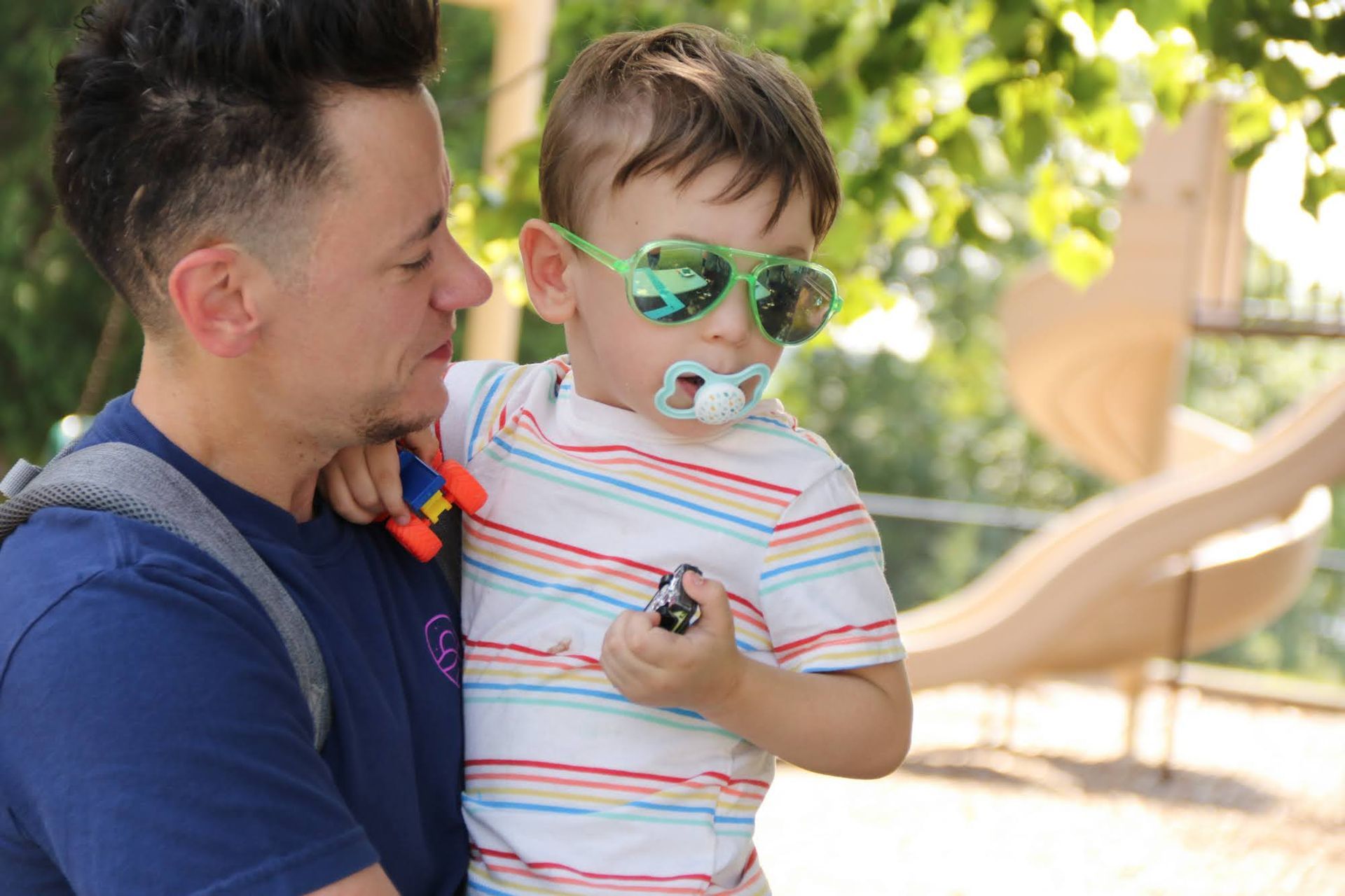 Man holding a child wearing sunglasses and a pacifier outdoors near a playground.