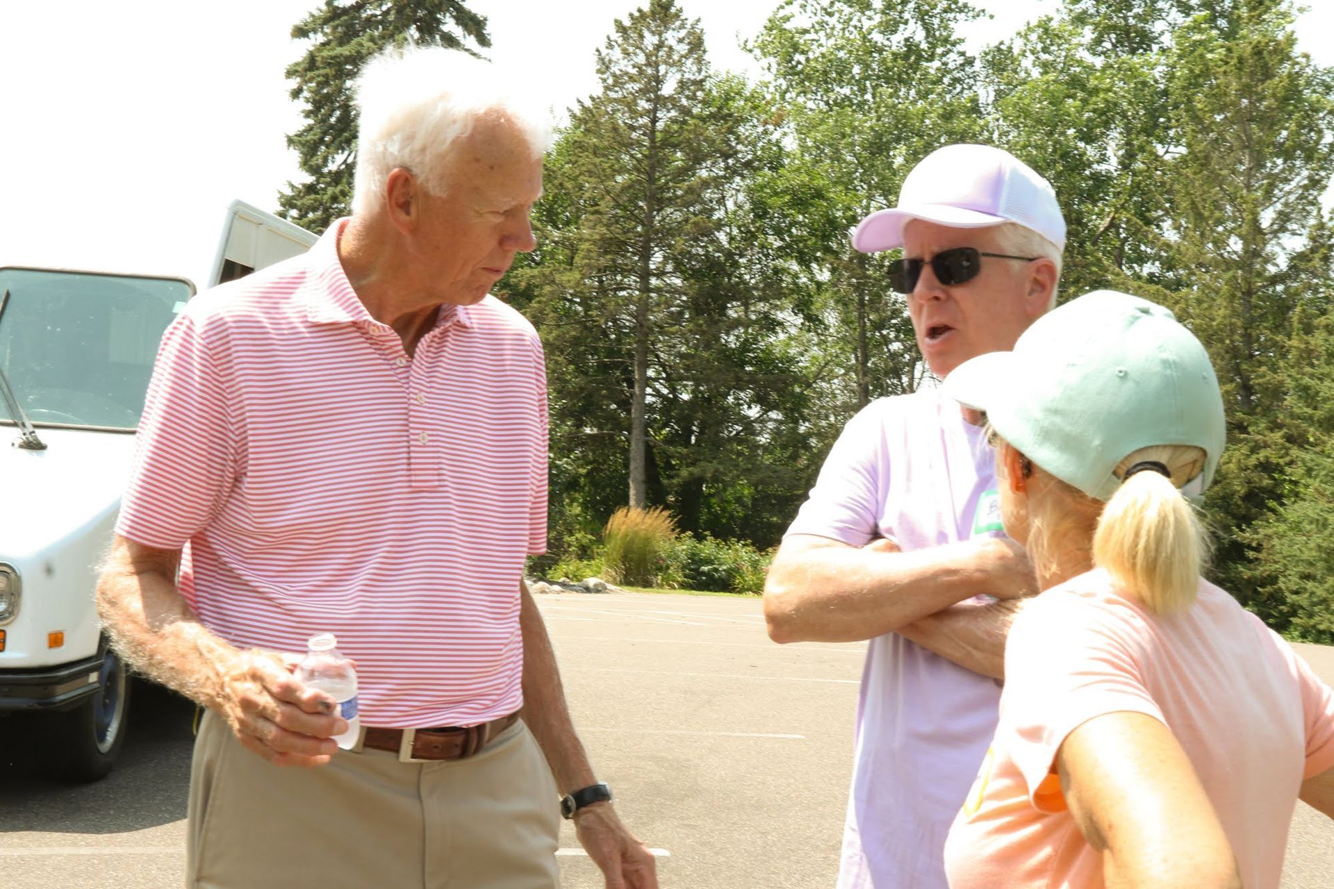 Three people standing in a parking lot. Two men in pink shirts converse while a woman with blonde hair listens.