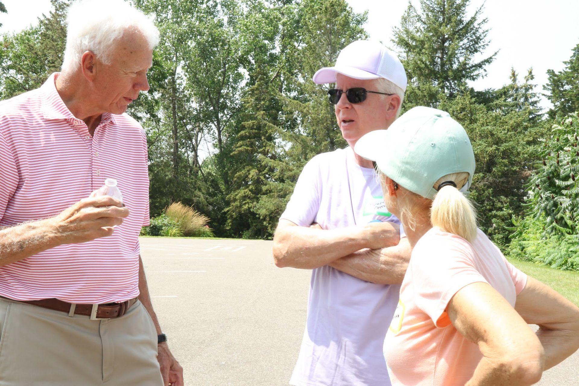 Three people in casual wear talking outdoors. One man holds a small object, the other has arms crossed. Trees in background.
