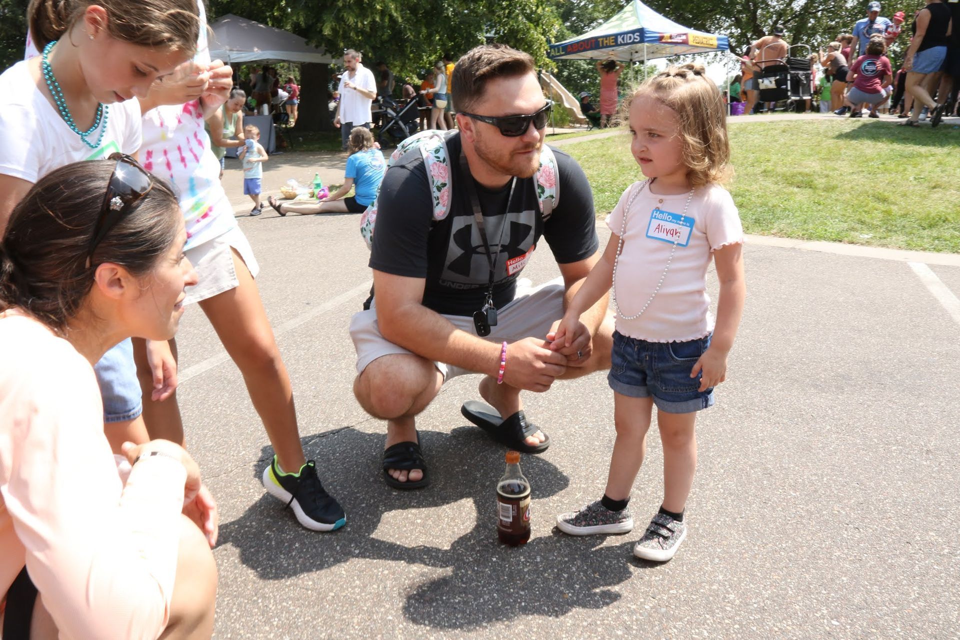 Man crouches, holding hands with a young girl outside. Two other people stand nearby. Sunny outdoor setting.
