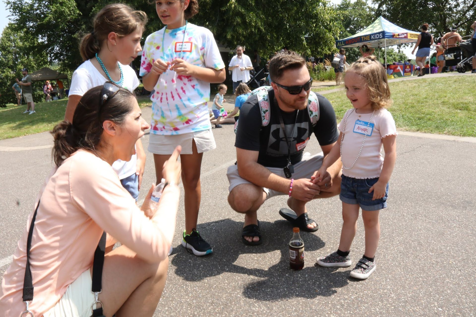 Group of people outdoors: two adults and three children by a soda bottle; sunny day.