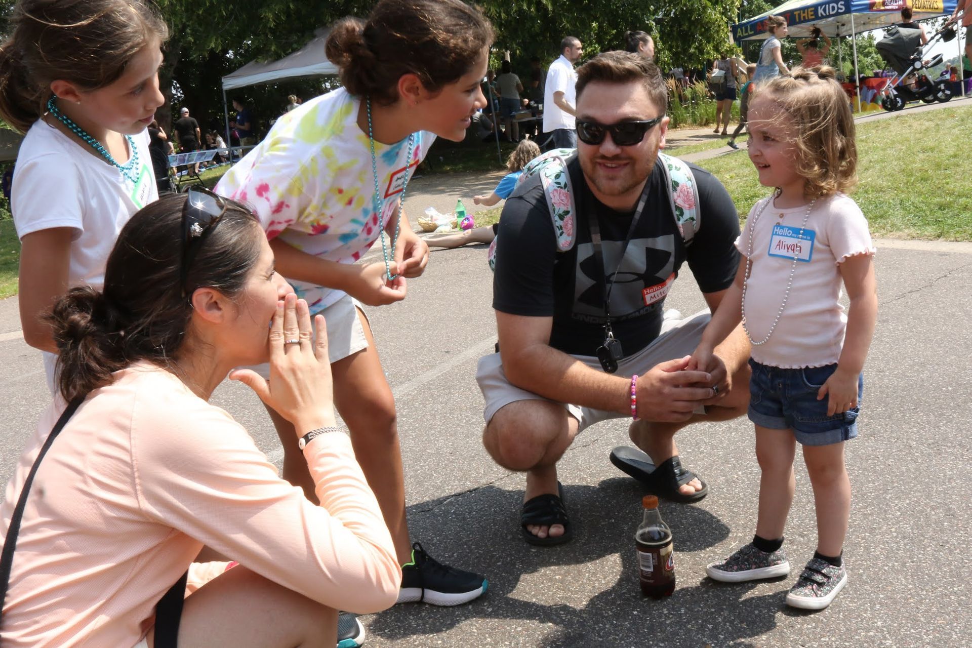 A group of people surround a bottle on the ground. A woman whispers to a girl, another girl looks on. A man crouches near a young child.