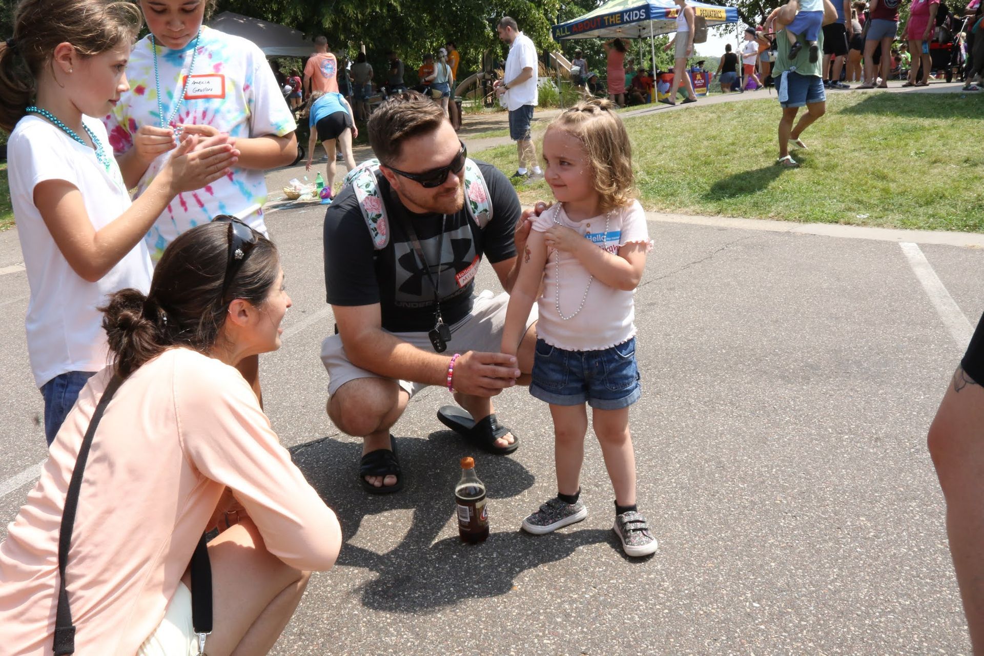 People gathered outside. Man kneels to speak to a girl, another girl and woman beside them. Bottle on the ground.