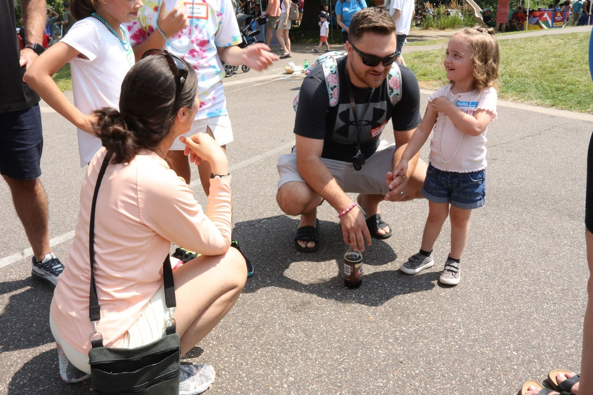 People gathered outdoors: A man and woman crouch, talking to a young girl. Other people and greenery are in the background.