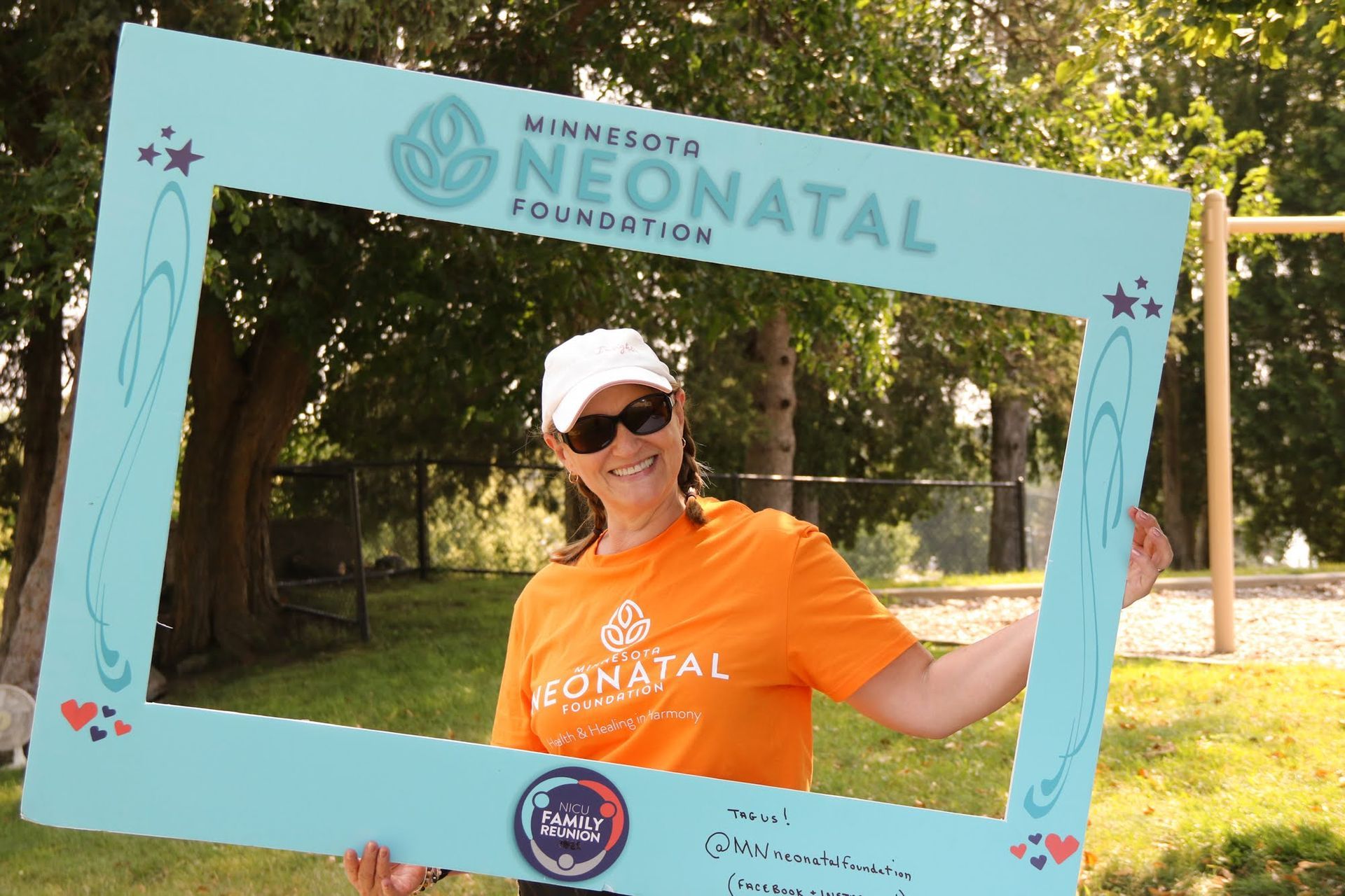 Woman in orange shirt poses behind a teal photo frame at an outdoor event. Frame has foundation logo.