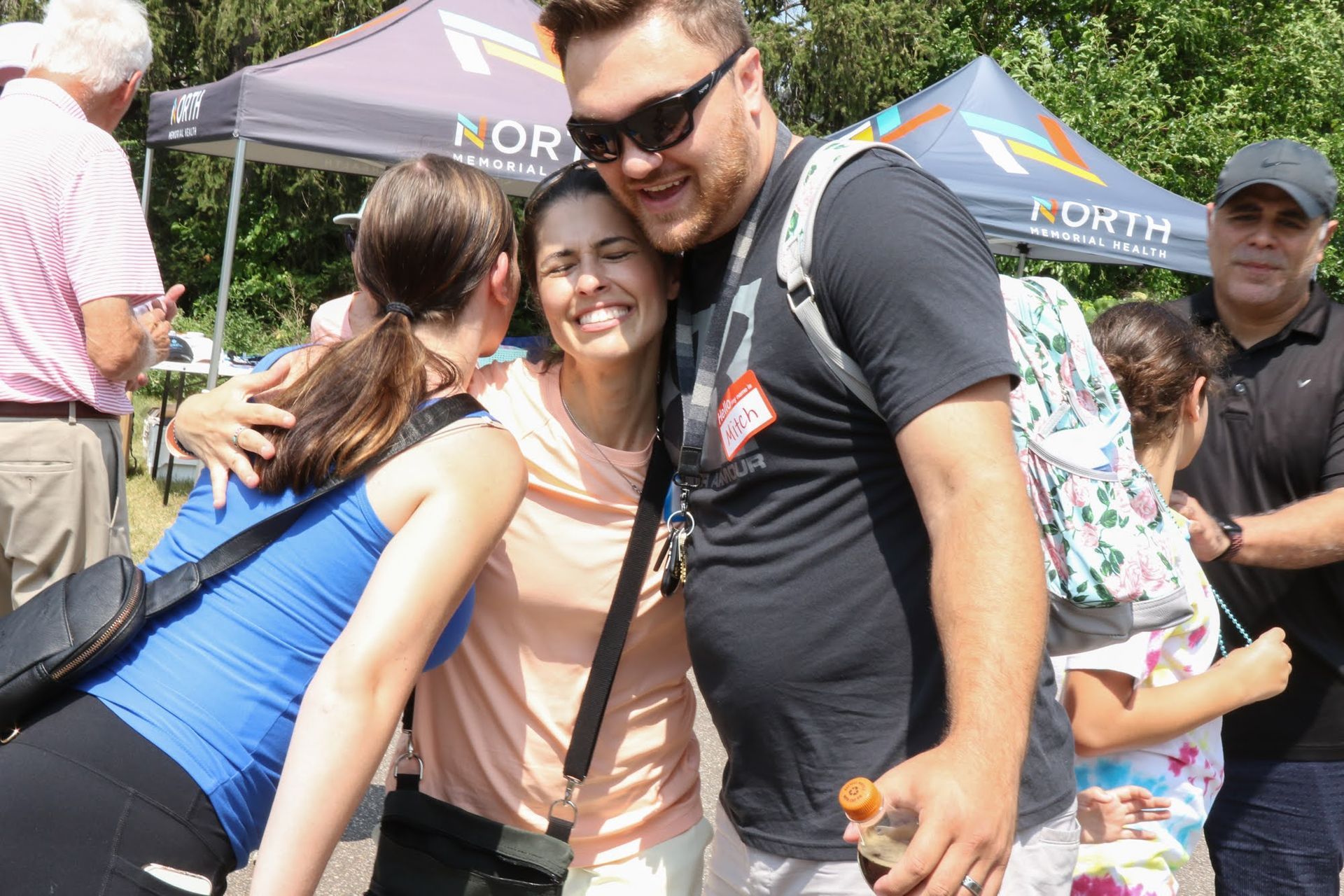 People embracing outdoors at an event, under tents. Smiling and wearing sunglasses.
