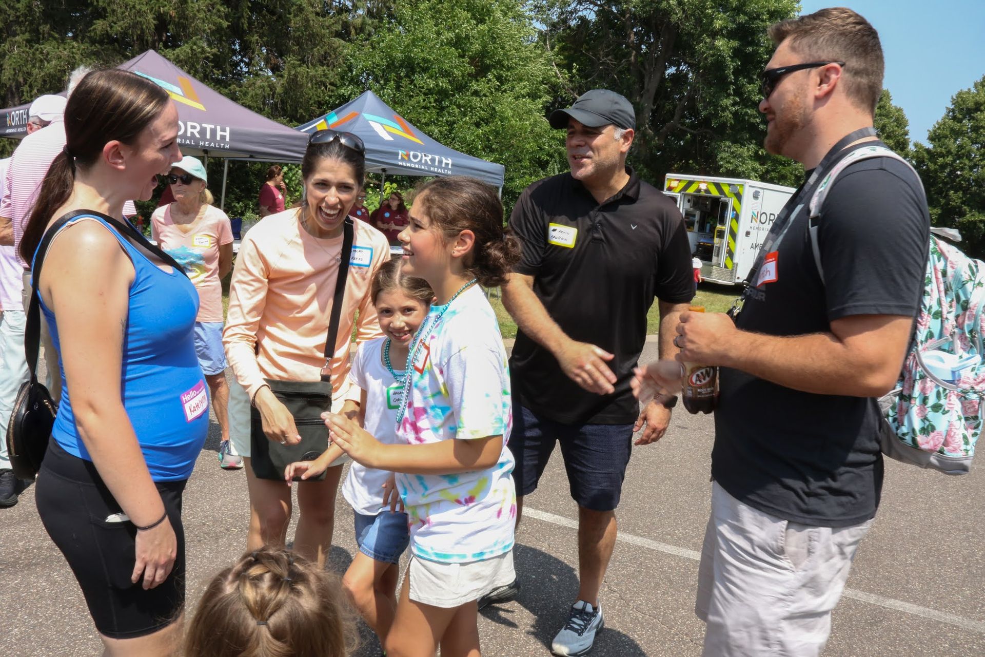 People smiling and talking outdoors at an event, surrounded by tents, trees, and parked cars.