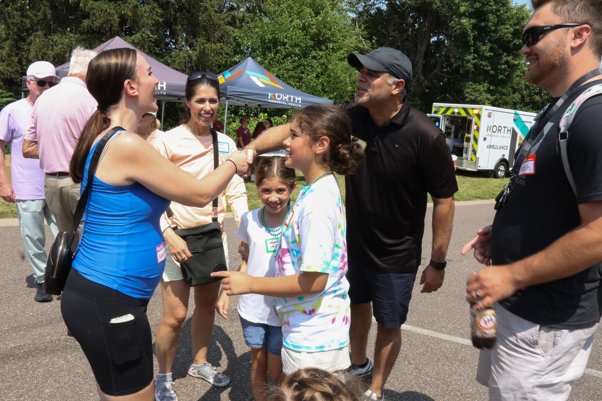 People greeting each other at an outdoor event; includes a child, adults, tents, and an ambulance.