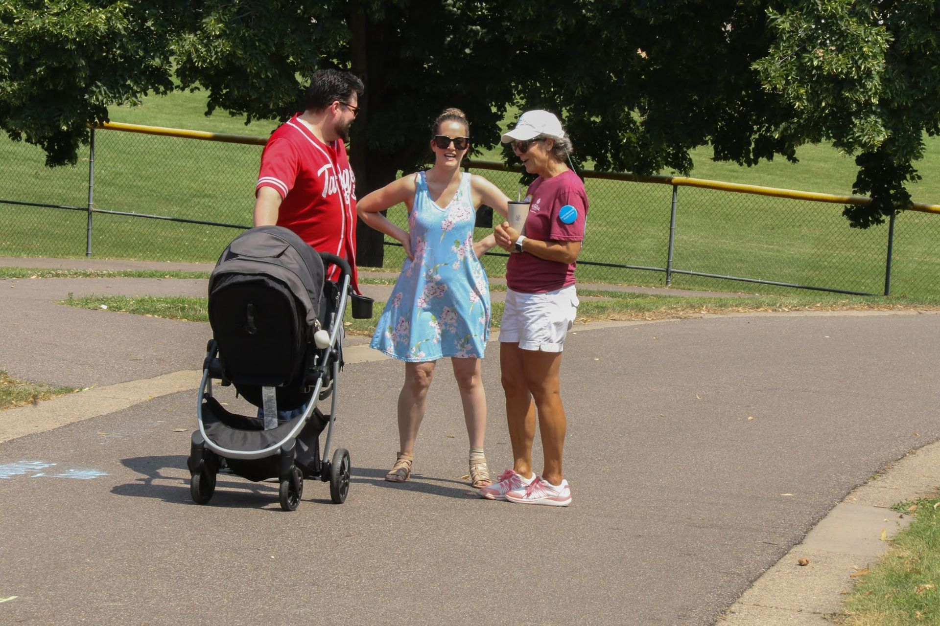 Three people chatting on a paved path in a park; a stroller is next to a man in a red shirt.