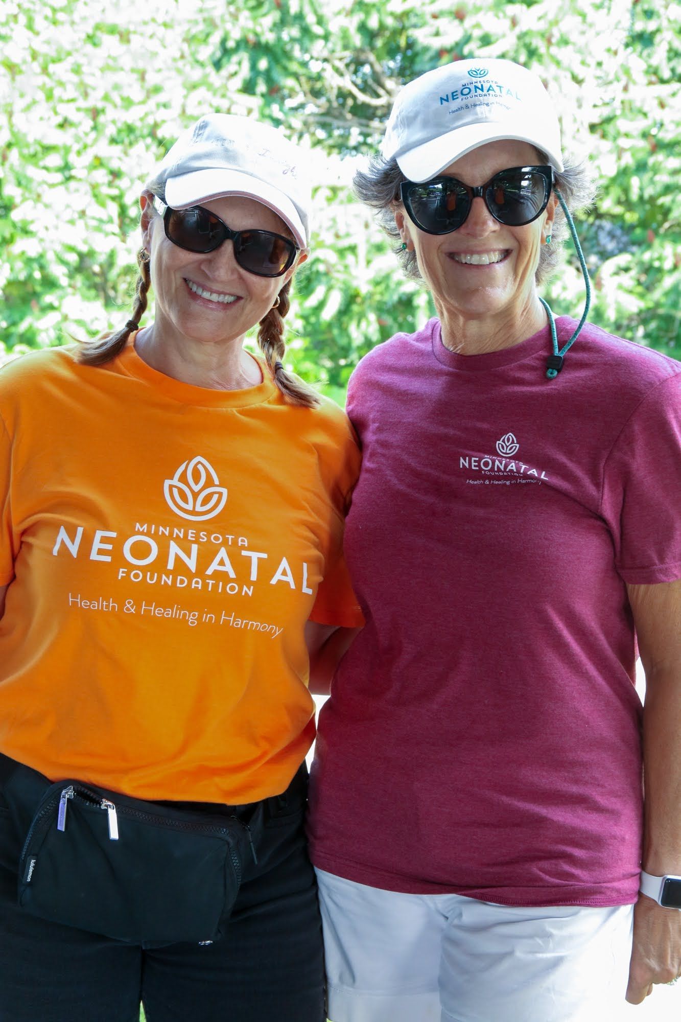 Two women in sunglasses and hats, smiling. One in orange shirt, the other in burgundy. Outdoor setting.