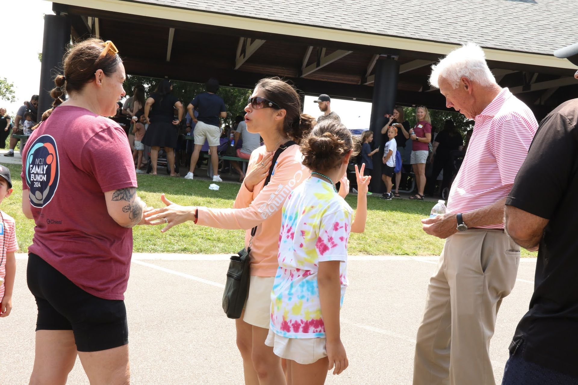 People talking outdoors, near a covered structure. One woman wears a tie-dye shirt, others wear casual clothing.