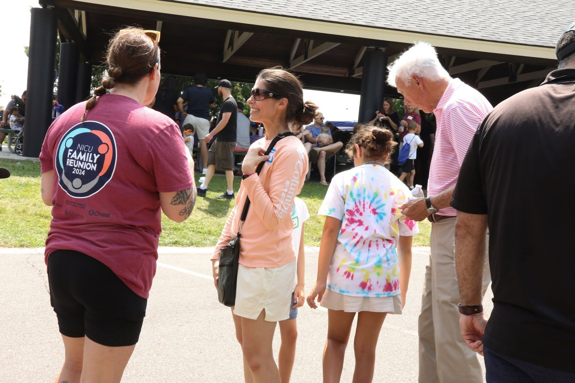 People gathered outside a pavilion. Woman in maroon shirt, another in peach, girl in tie-dye.
