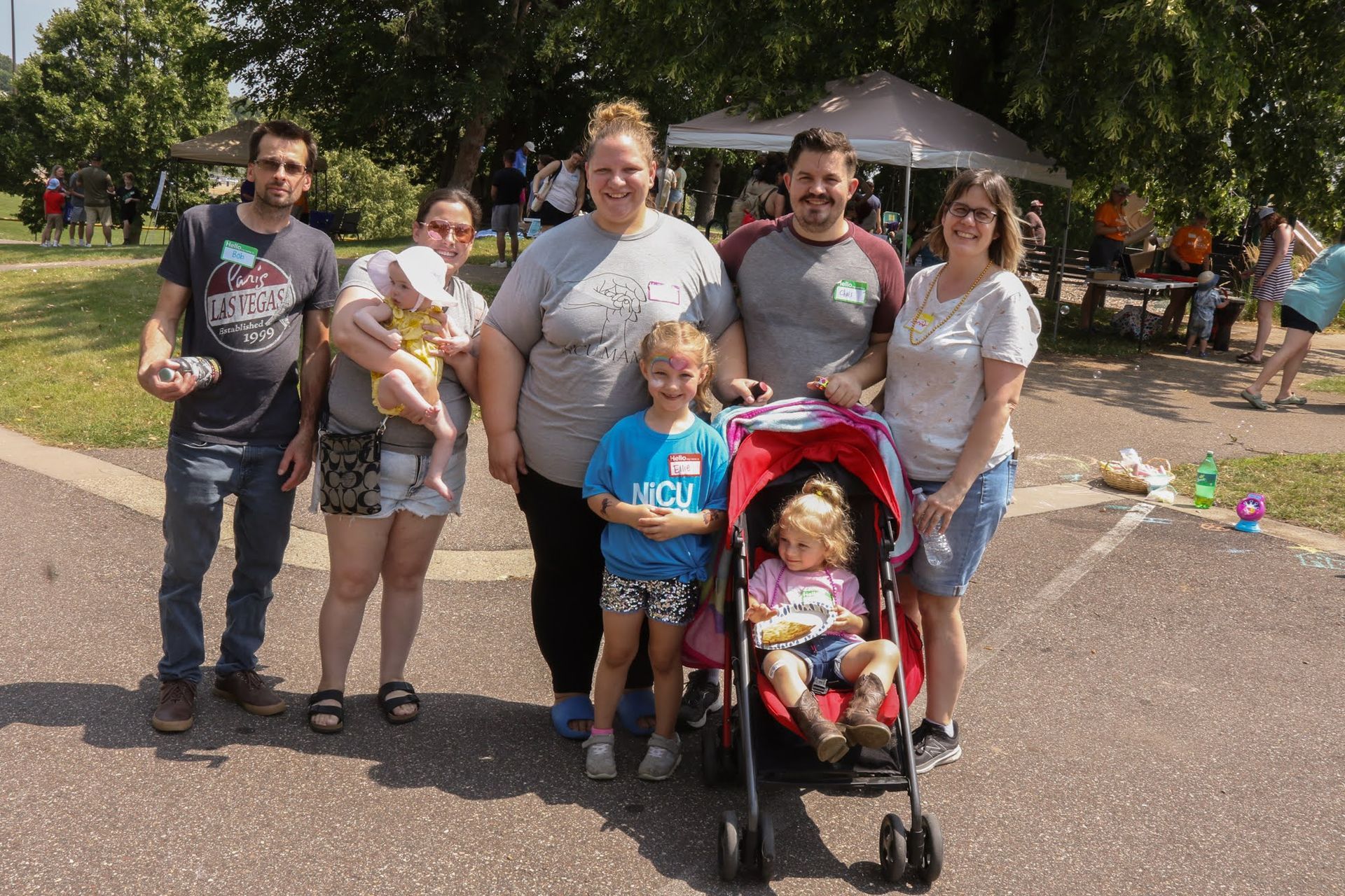 Group of seven people smiling for a photo in a park, two children in a stroller and held.