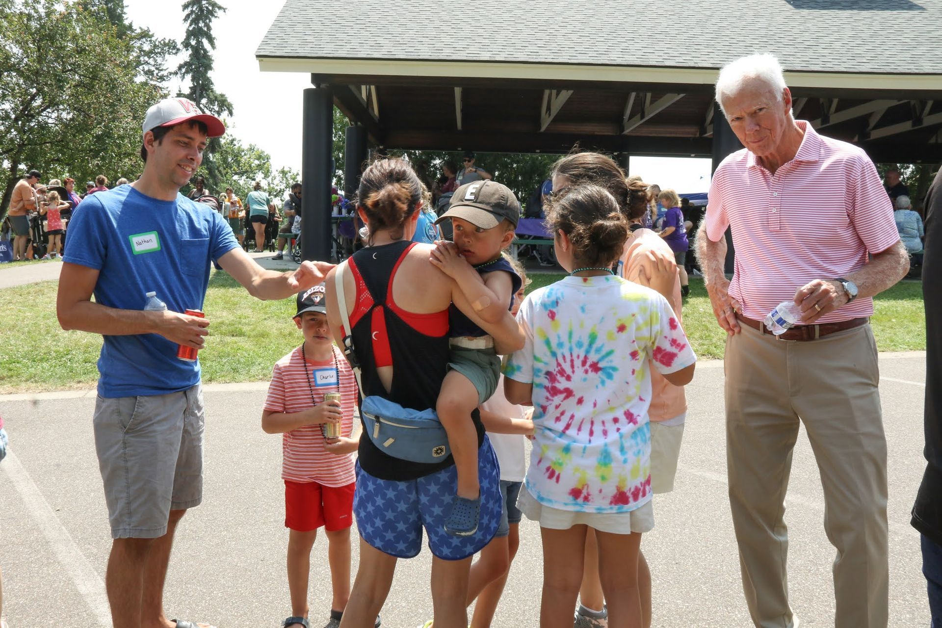 People at an outdoor gathering: a man in a pink shirt, children, and a woman holding a child.