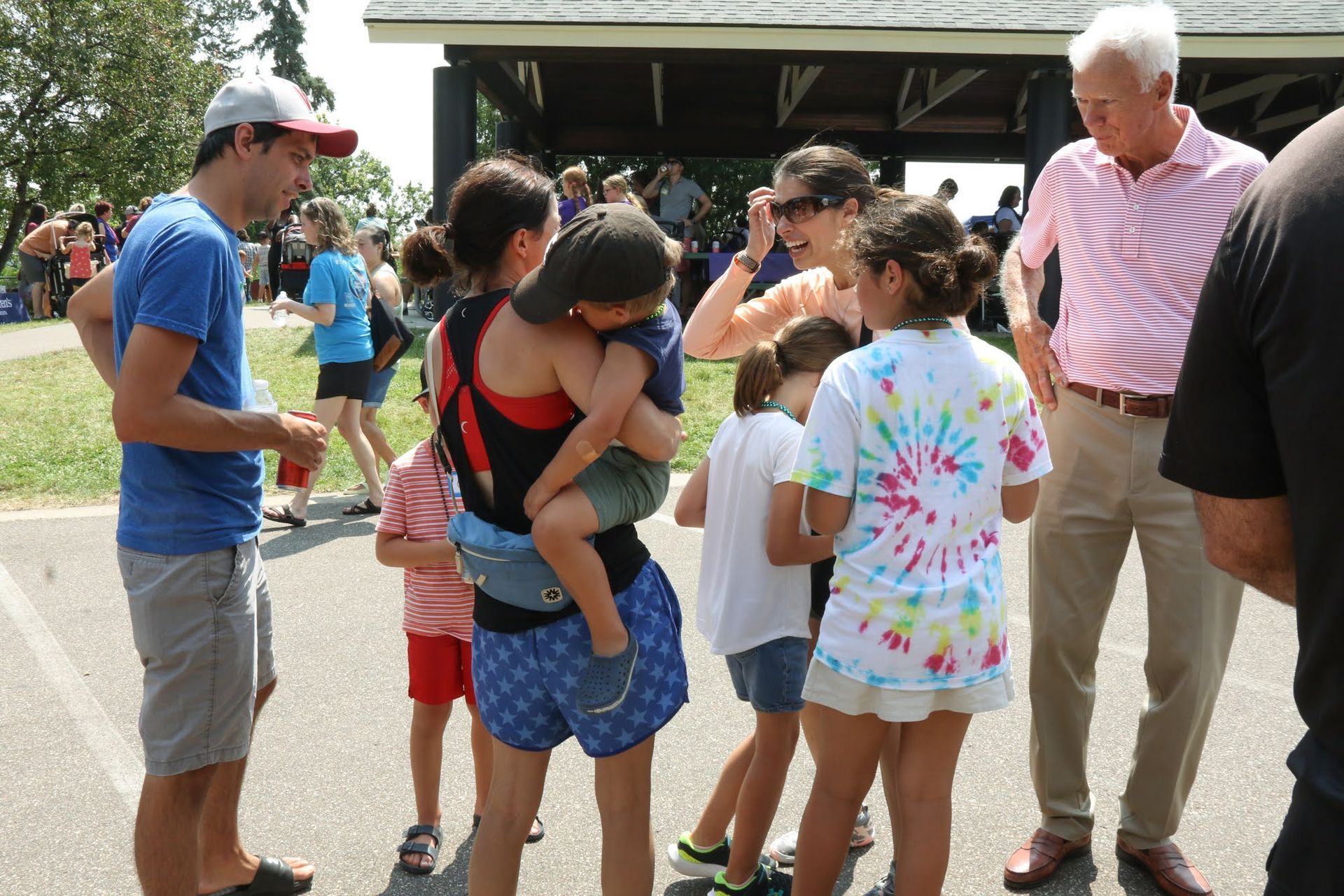 People gather near a pavilion. One person carries a child. Others converse. Sunny outdoor setting.