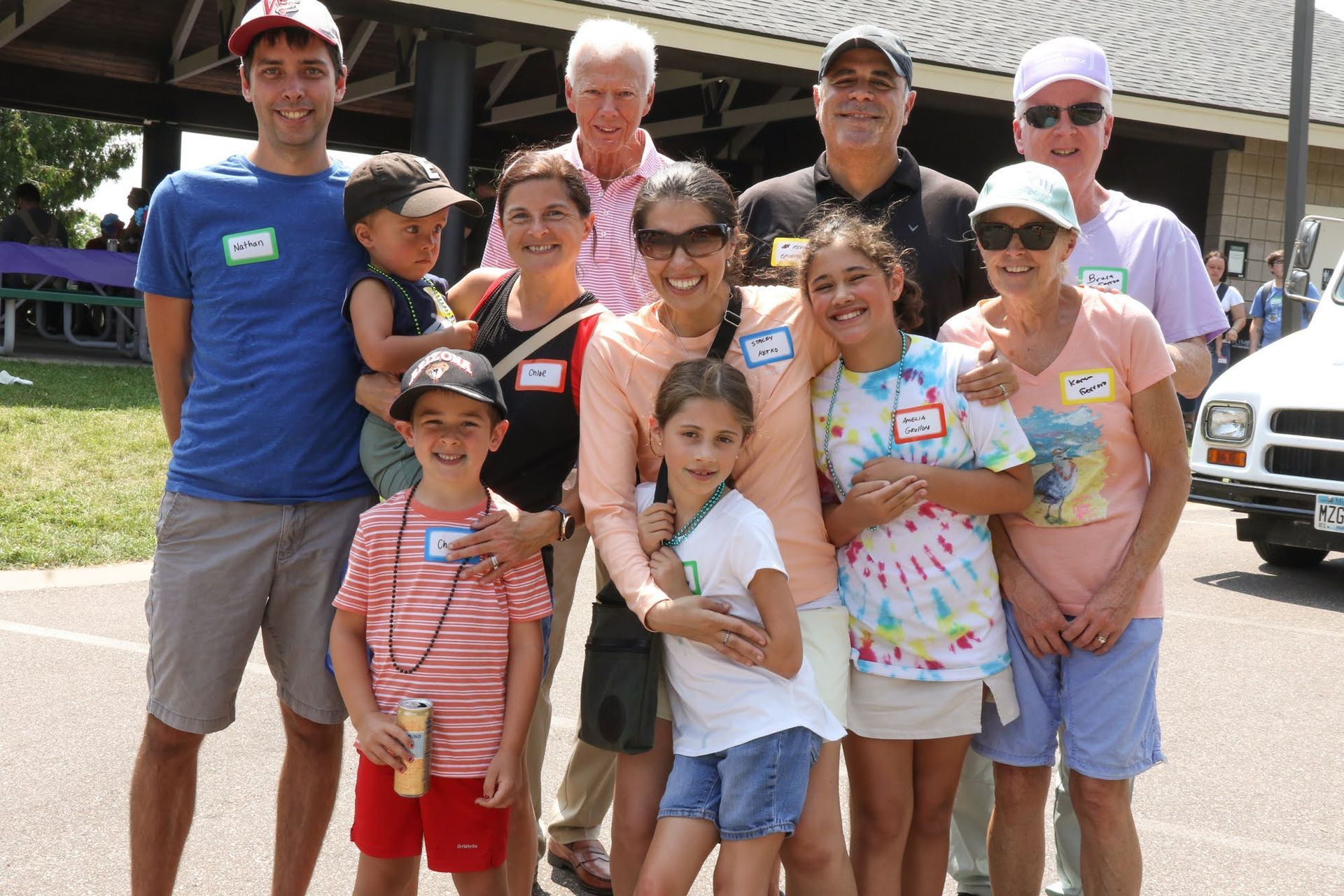 Group of people posing outdoors, smiling. Some children in front, adults behind, near a pavilion.
