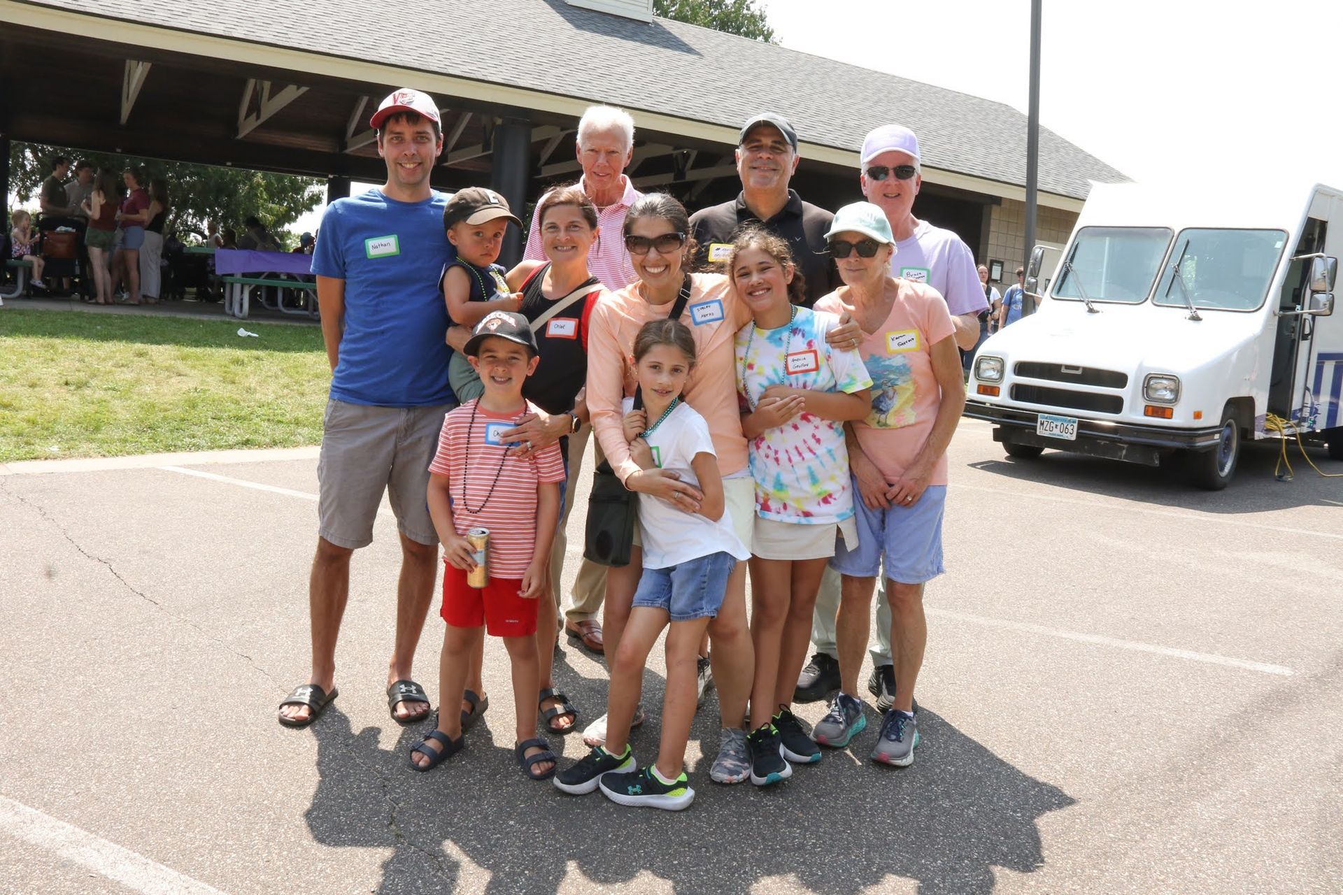 Group of people posing outdoors near a white van and pavilion on a sunny day.