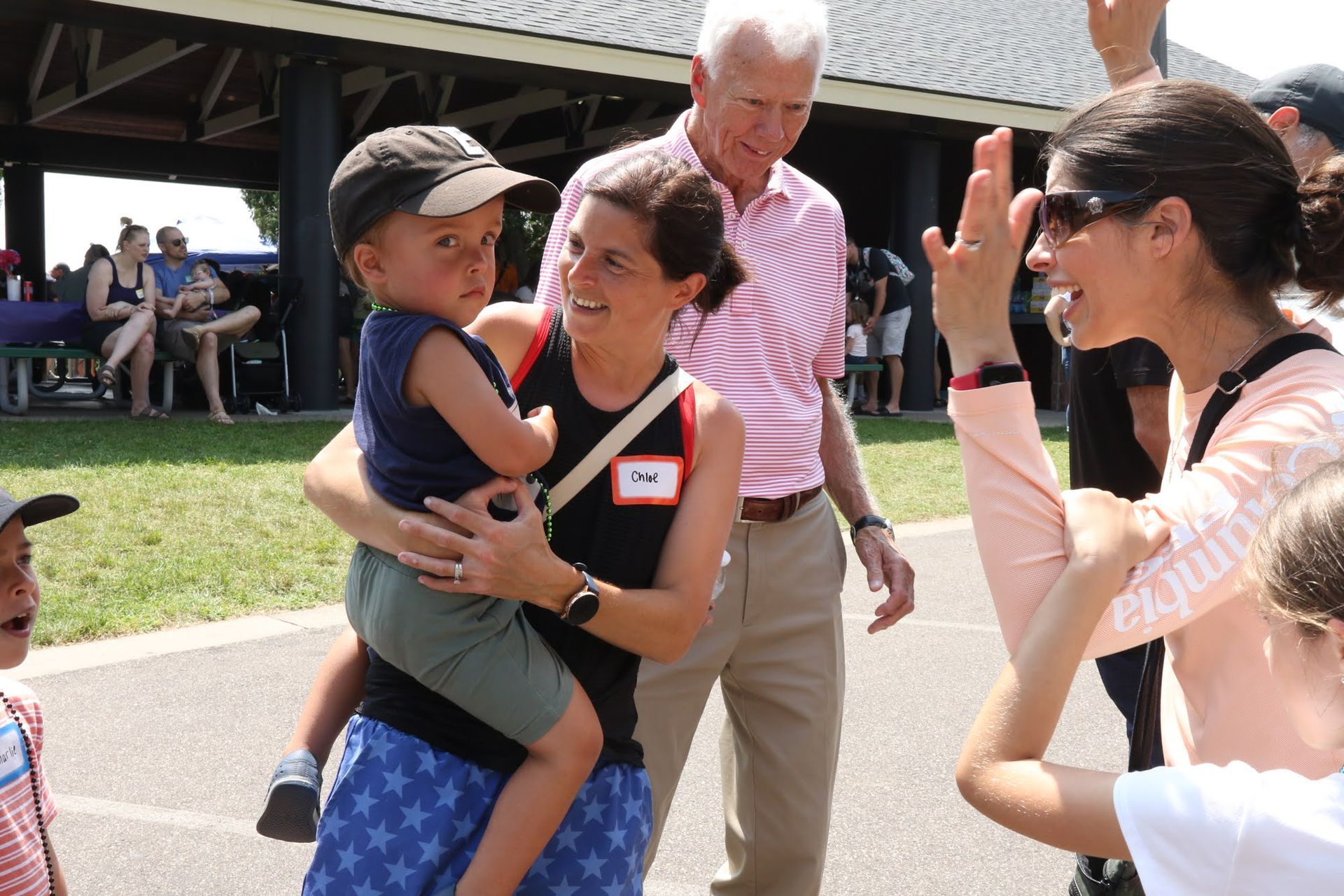 Woman holding child smiles at another woman giving a high five, park setting.