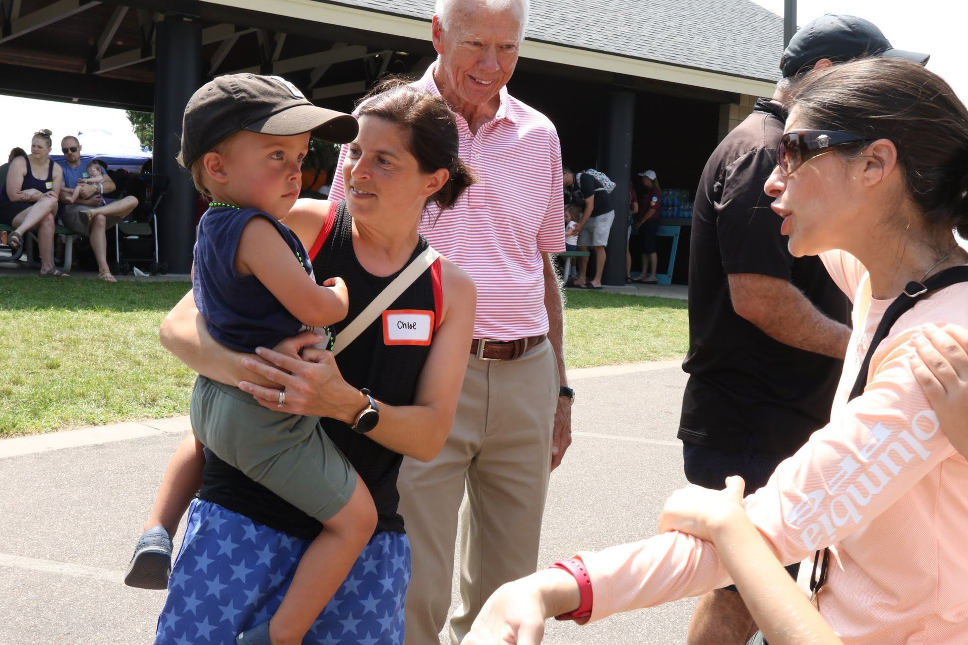 Woman holding child, talking to another woman outside. Man in pink shirt nearby.