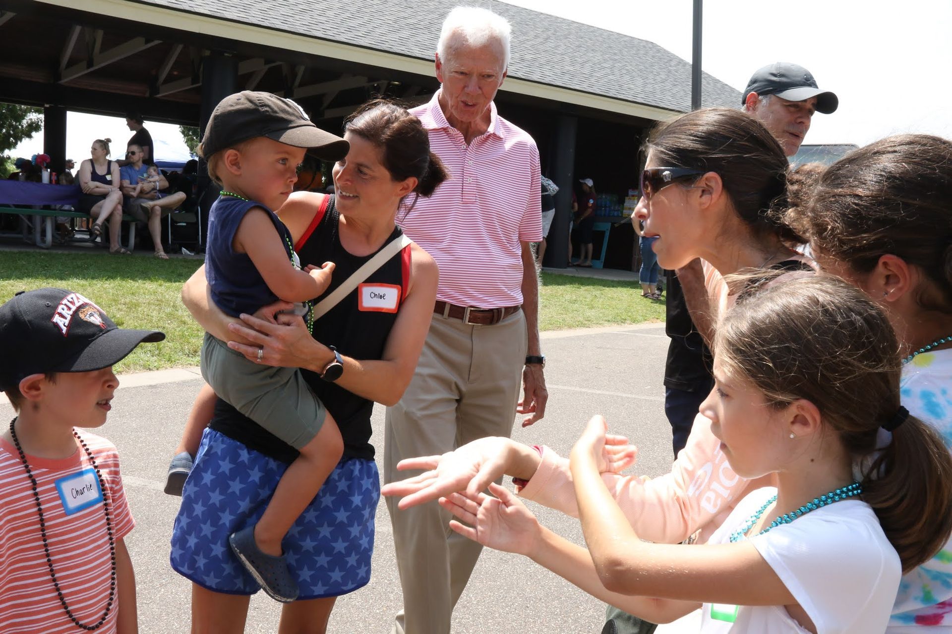 People interacting outdoors: a woman holds a child, others talk and gesture in a park setting.