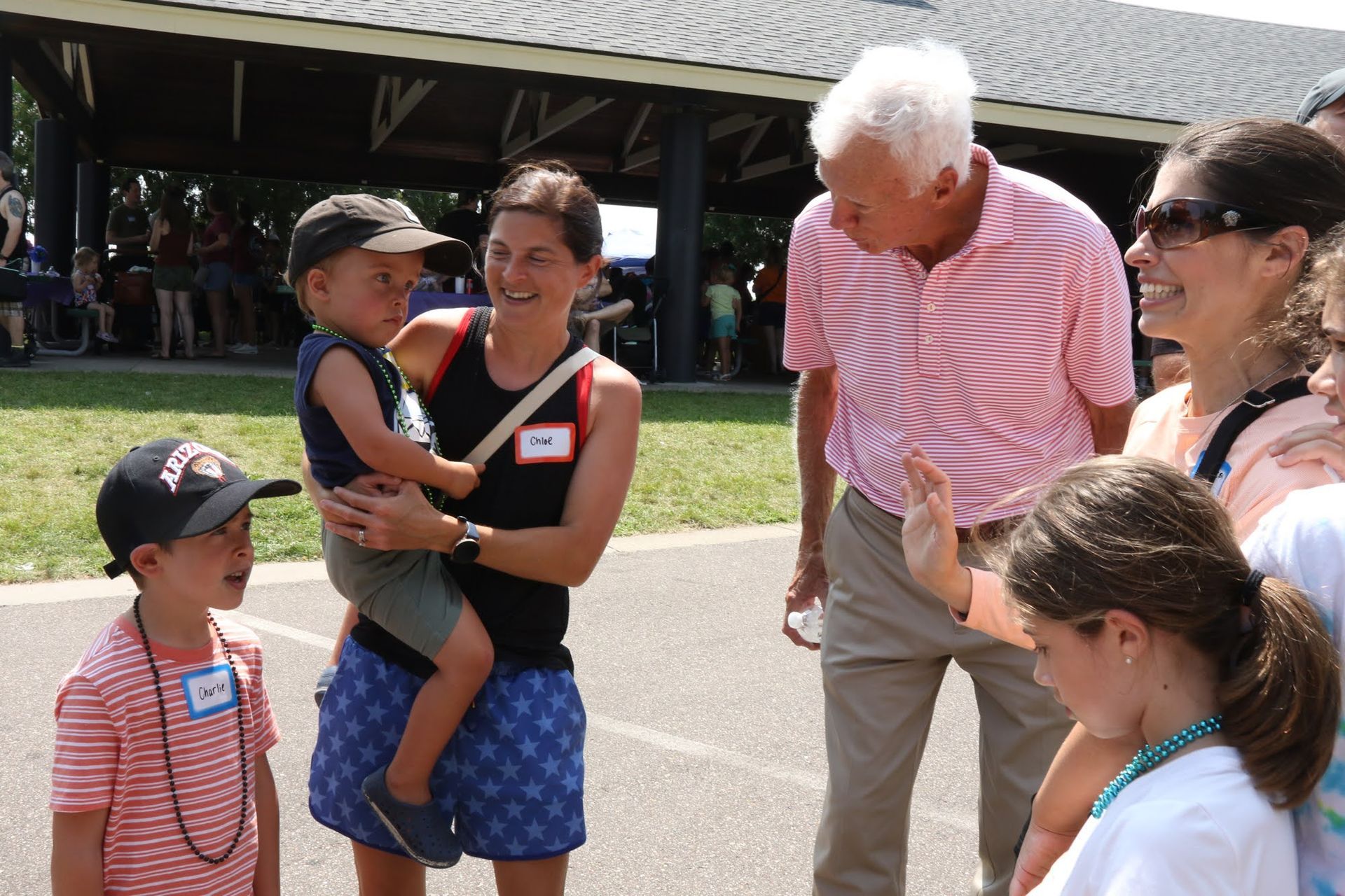 A man speaks to a woman holding a child; other children and adults look on, outdoors.