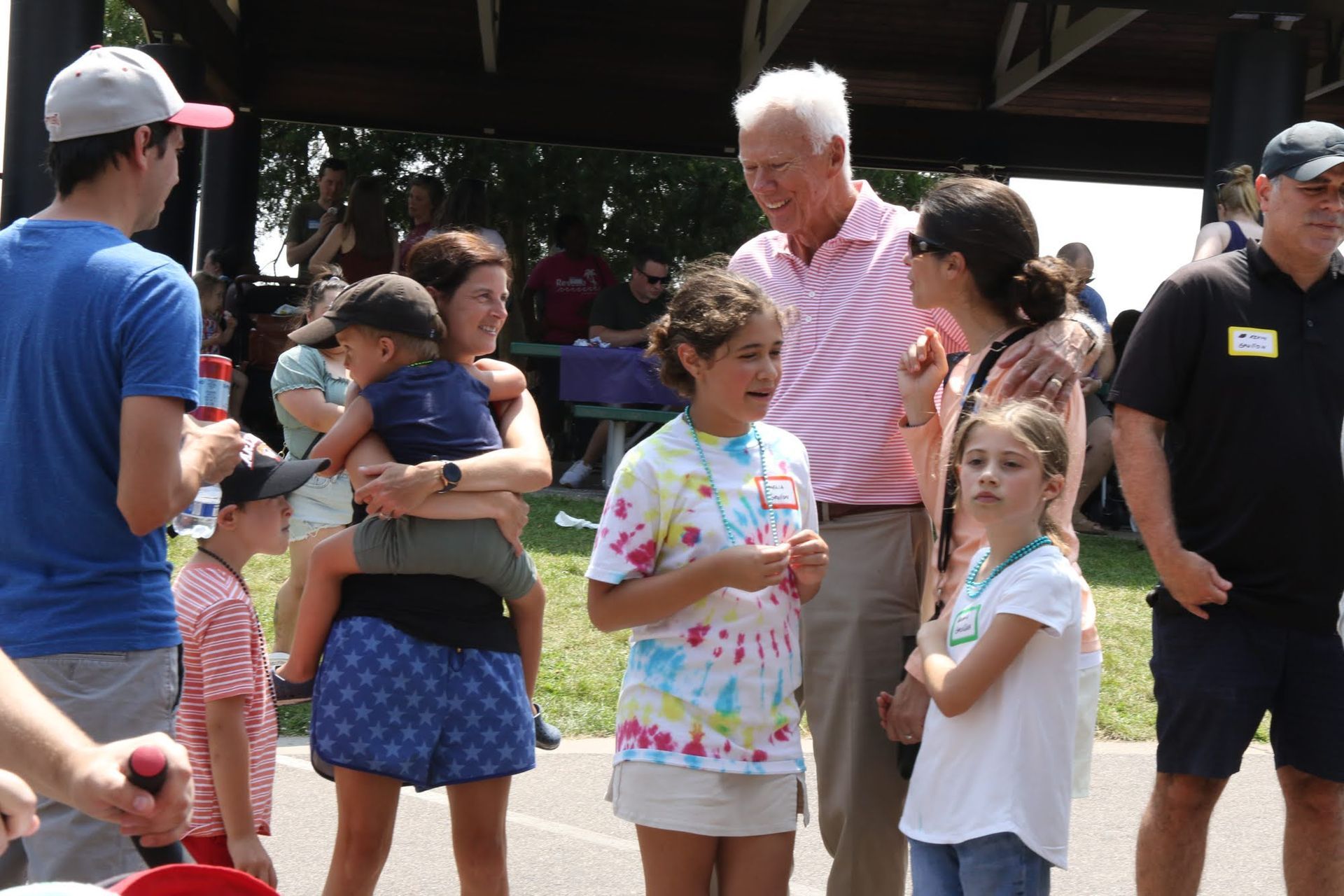 People at an outdoor gathering, including children, interacting with an older man wearing a pink striped shirt.