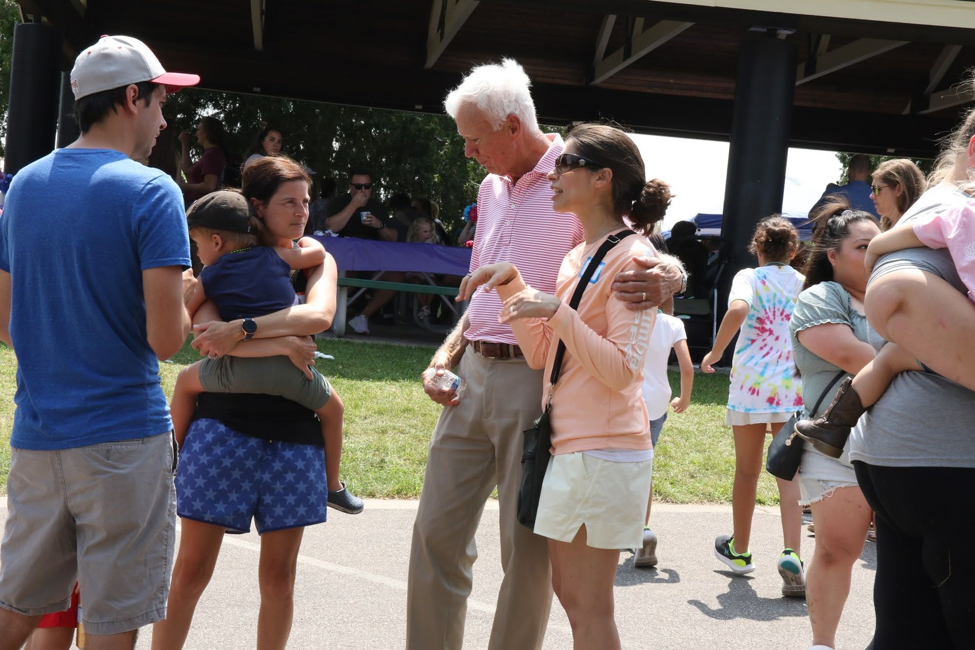 People outdoors, some interacting, under a pavilion. One man in pink shirt gestures while speaking. Others nearby.