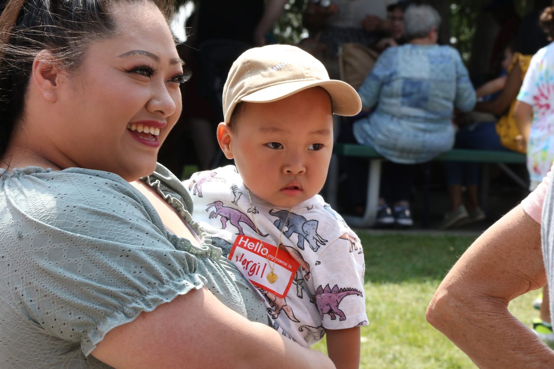 Woman smiles, holding a young child wearing a hat and dinosaur shirt at an outdoor gathering.