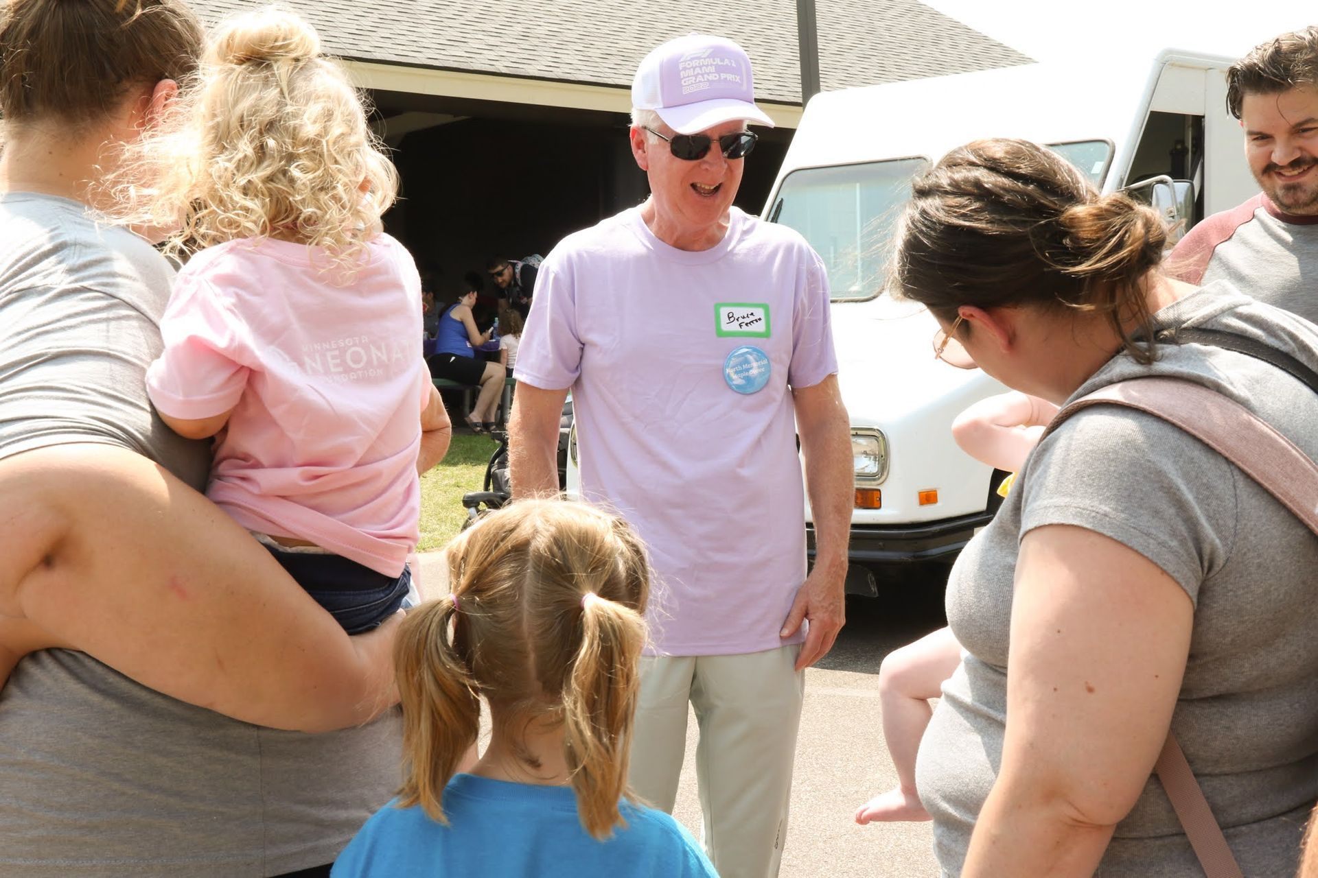 Man in purple shirt and hat speaks to people outside a van. Children and adults are present.