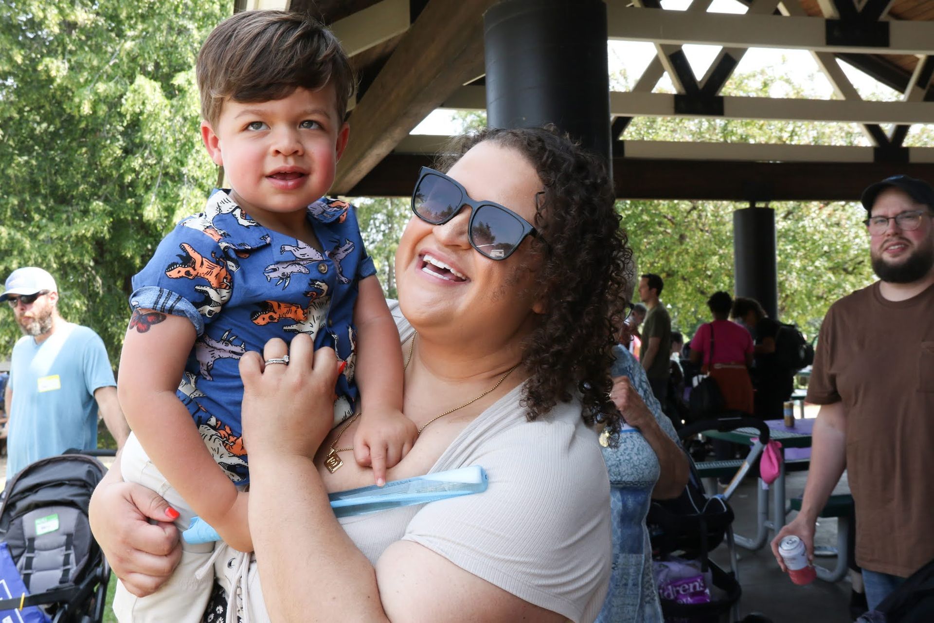 Woman holding a child smiling under a park shelter. People in the background. Sunny day.