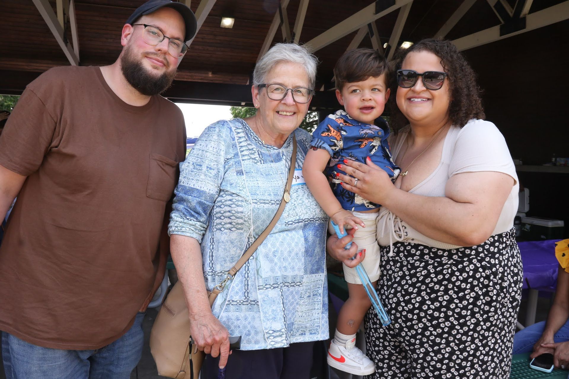 Family poses outdoors: man, woman, toddler held by another woman, smiling near a shelter.
