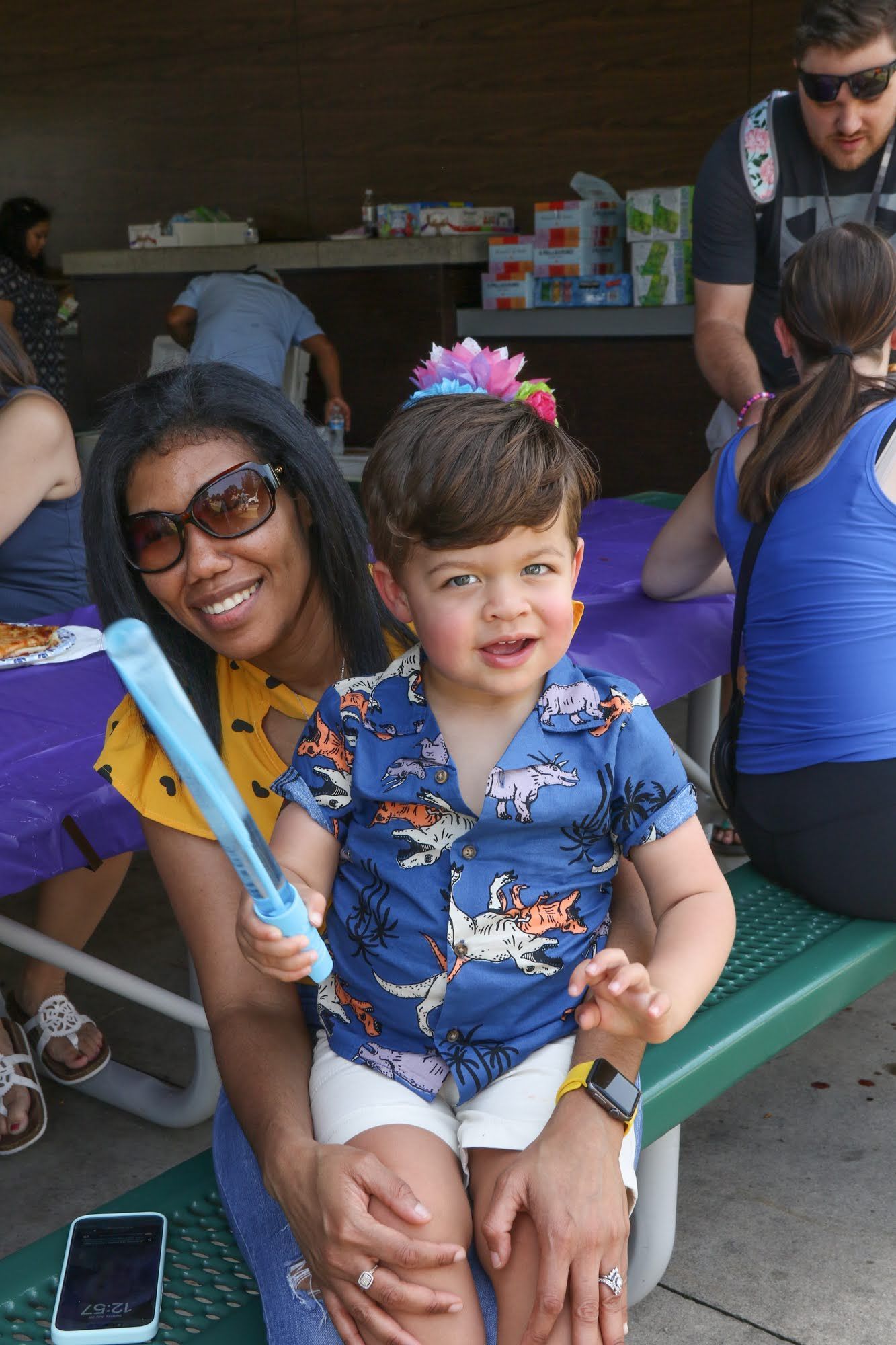 Woman with sunglasses holds a smiling child wearing a blue shirt and white shorts. Both are outdoors.