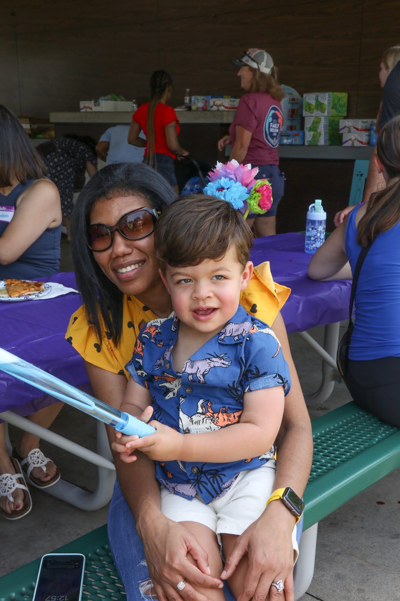 Woman and toddler smile while seated at a picnic table. Others are visible in the background.