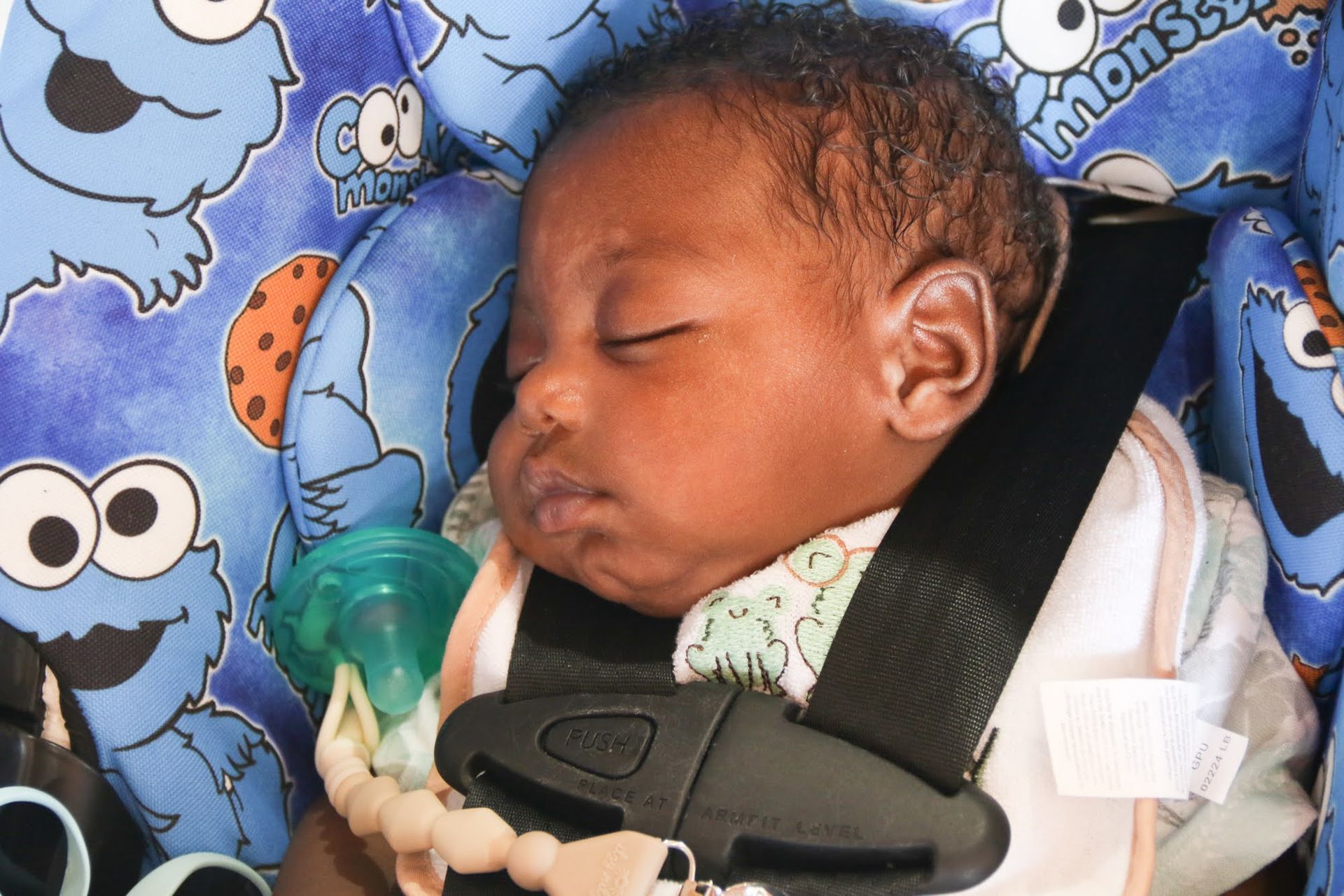 Baby asleep in a blue car seat with Cookie Monster print; pacifier visible.