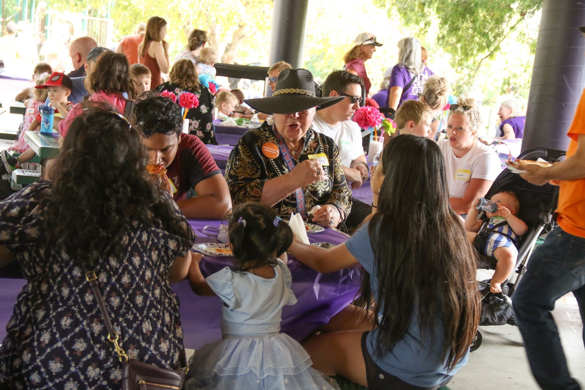 People eating at tables outdoors under a pavilion. A person in a hat is eating.