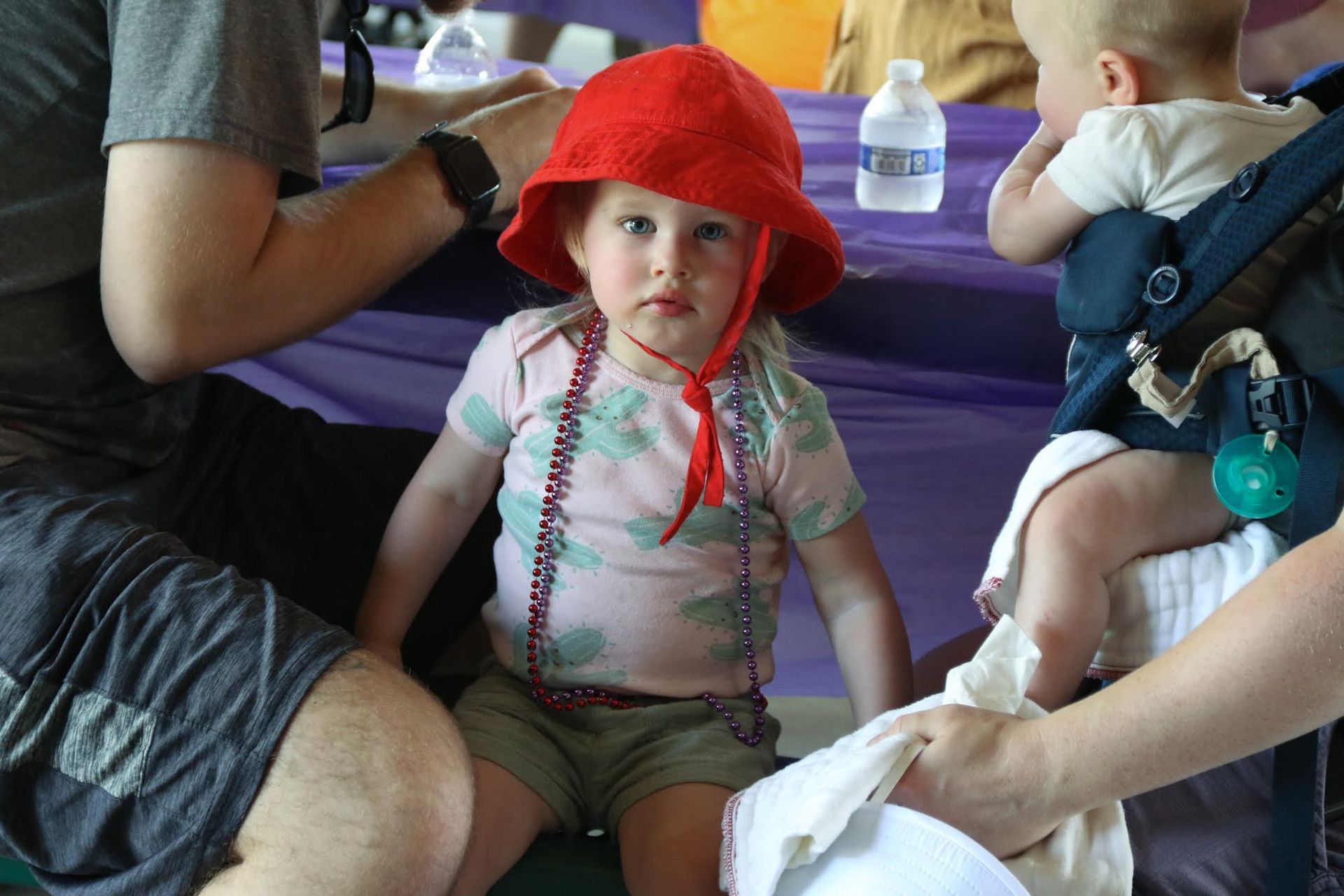 Girl wearing red hat and beads, sitting, looking at the camera.