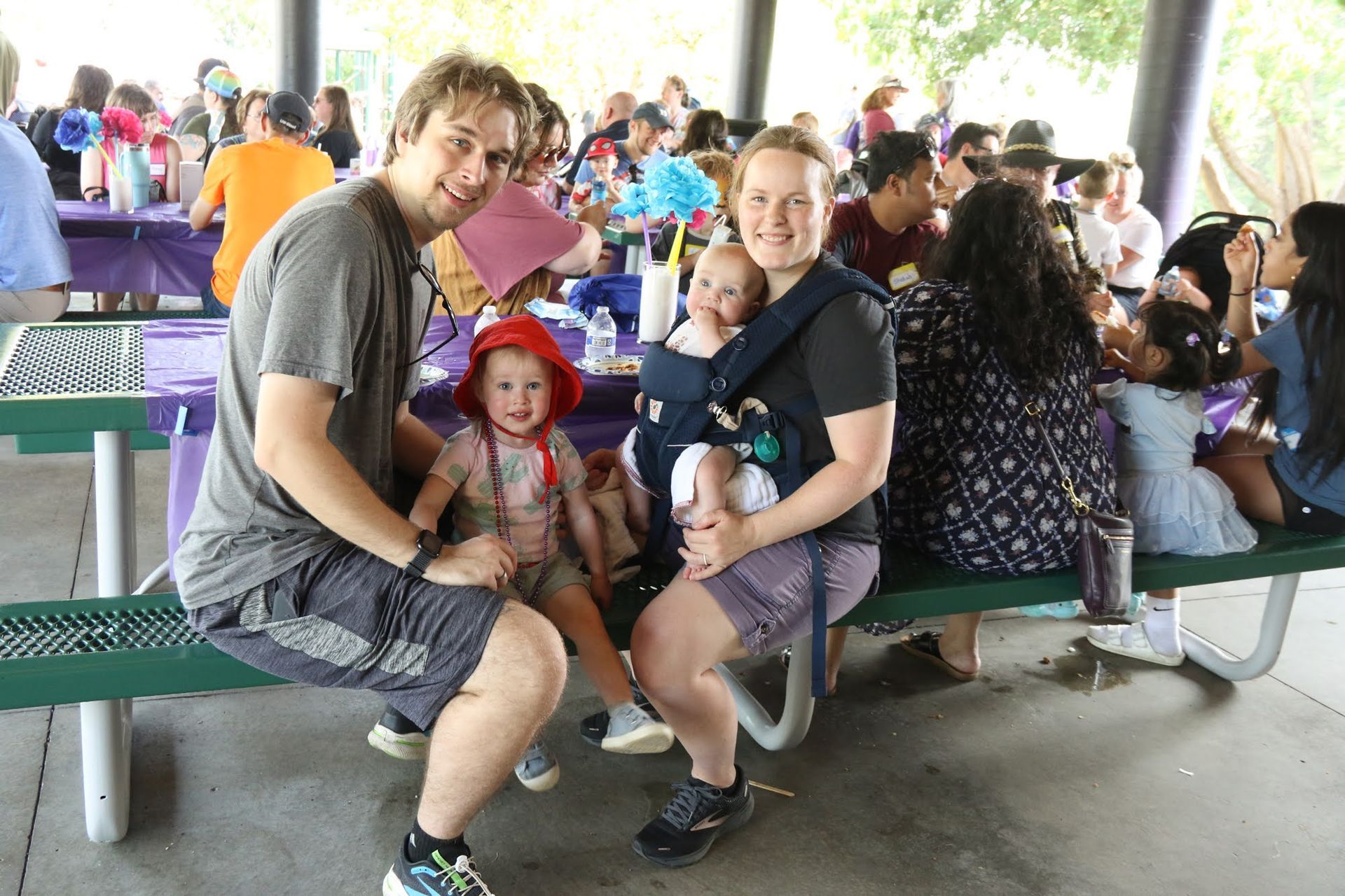Family of four at a picnic table: parents smiling, two young children. People in background, park setting.