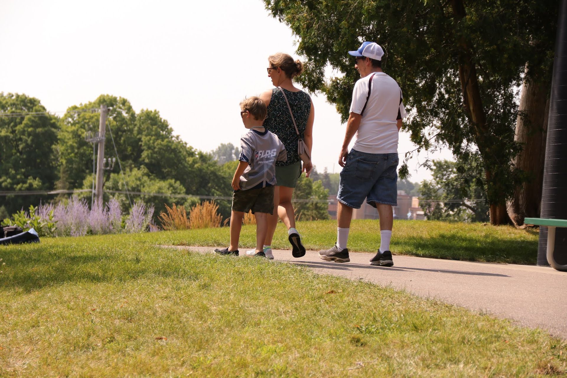 Family walking on a park path; trees and buildings in the background. Sunny day.