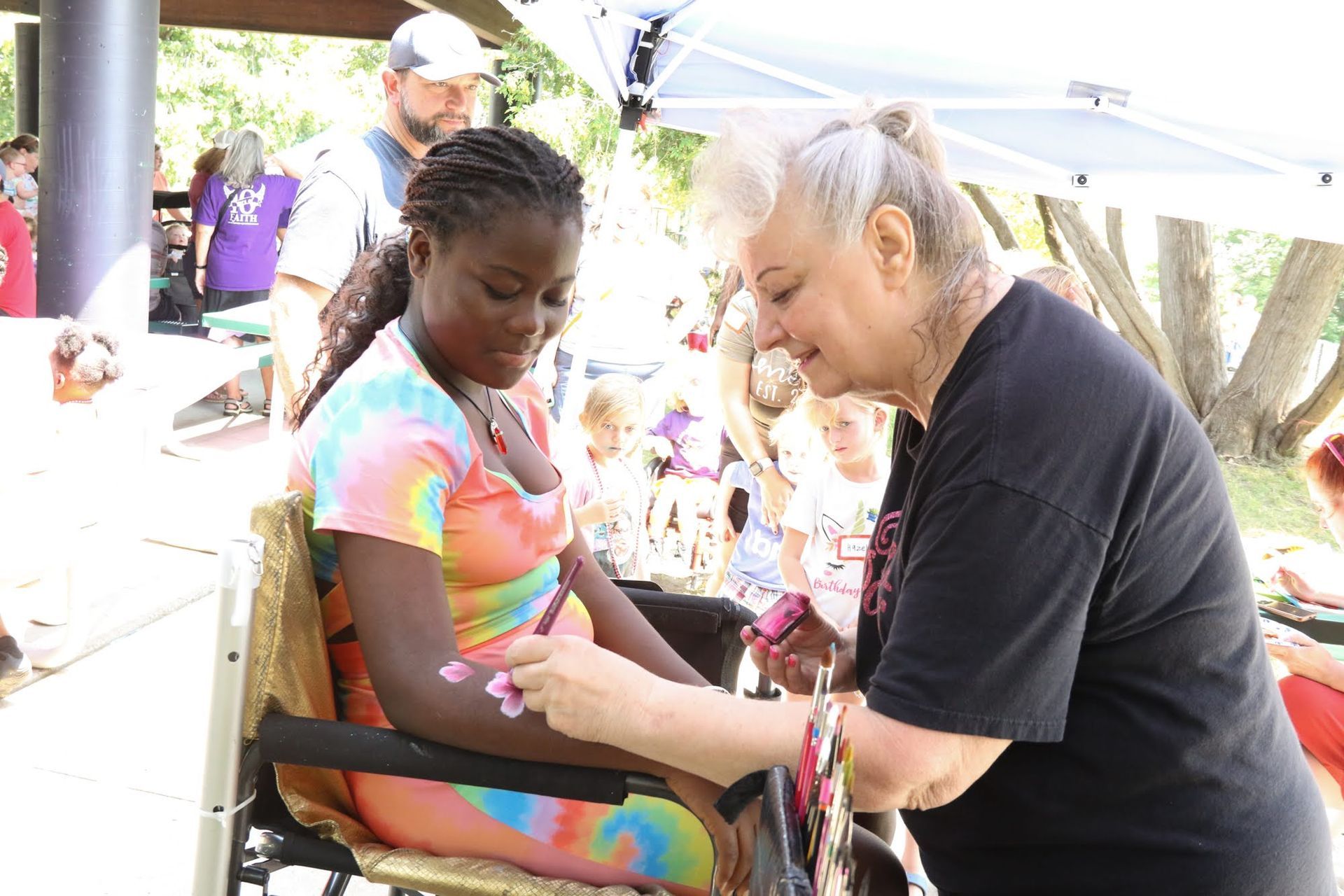Woman paints on the arm of a person in a wheelchair. Colorful event, outdoor setting.