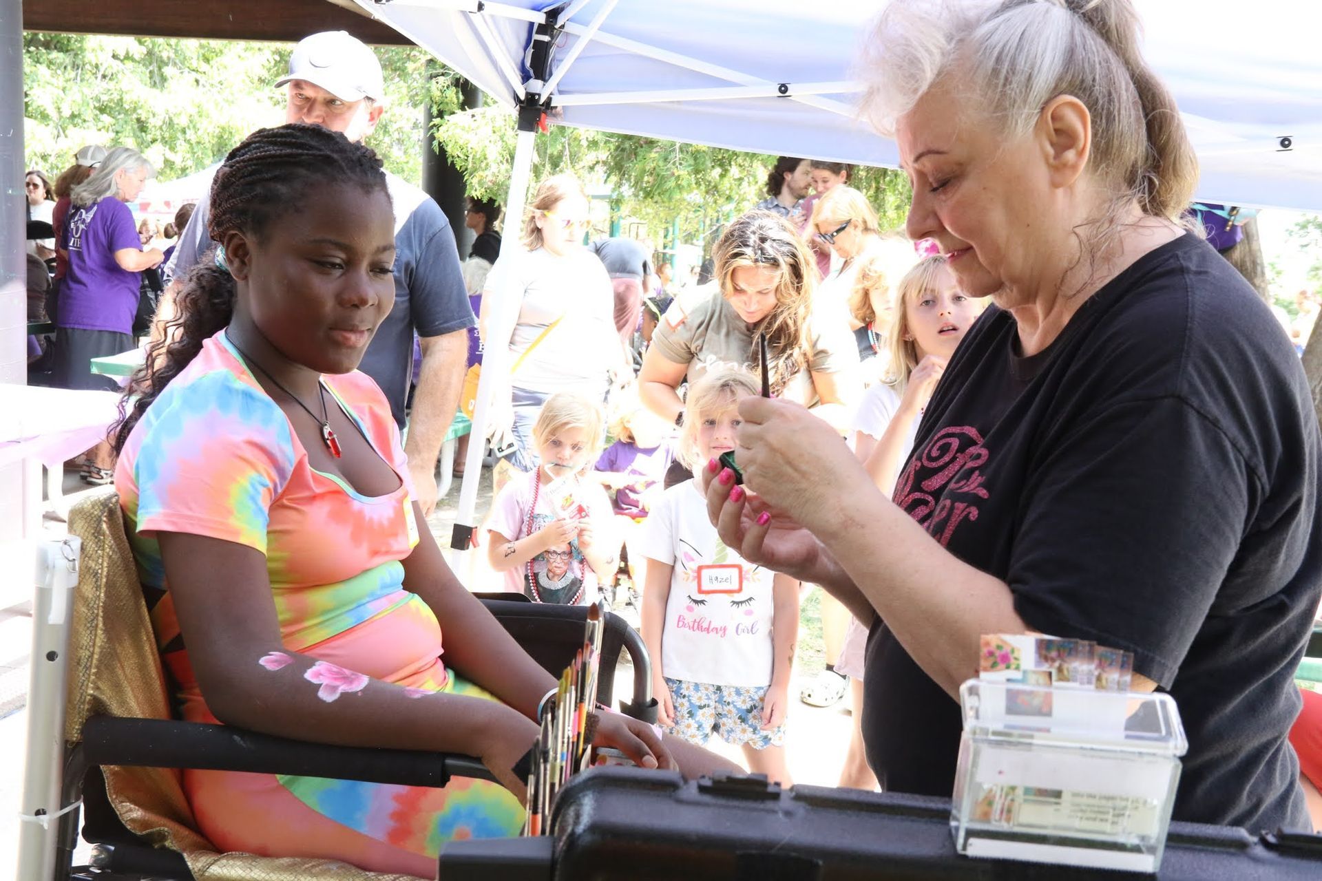Woman paints design on girl's arm at an outdoor event, surrounded by onlookers and a canopy.