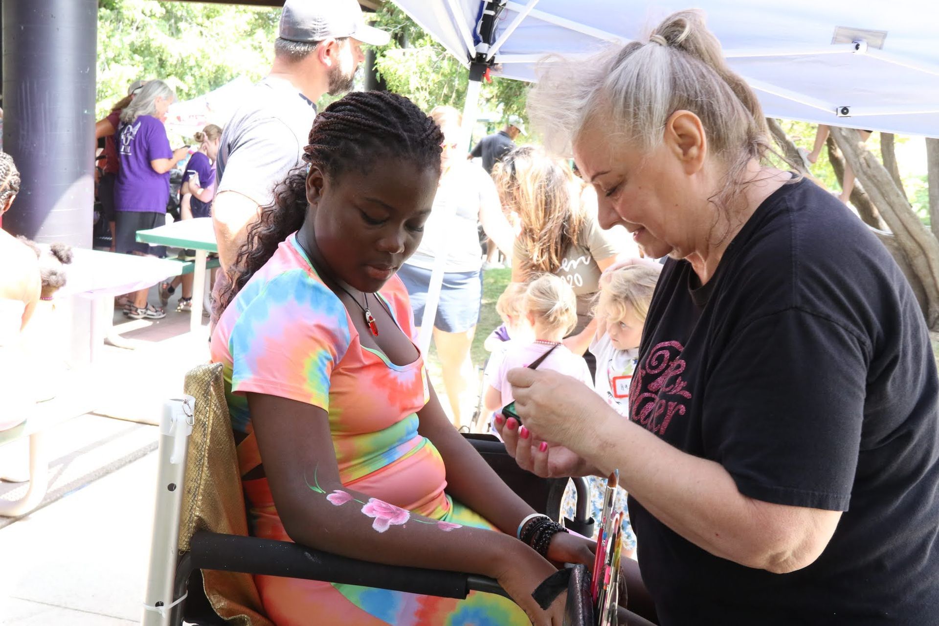 Woman paints flowers on the arm of a person in a wheelchair, outside at an event.