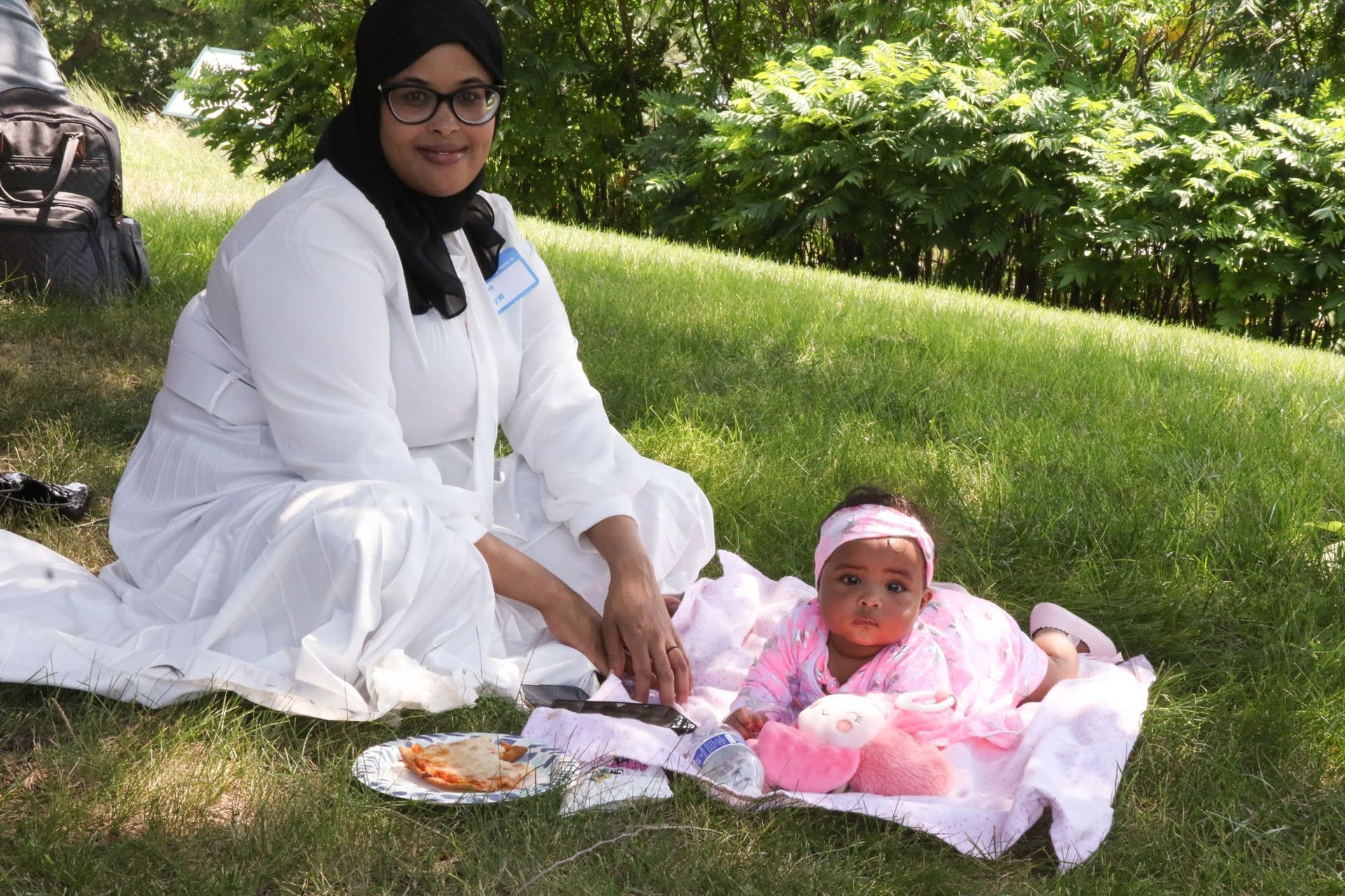 Woman in white attire sits on grass next to a baby in pink, both looking at the camera.