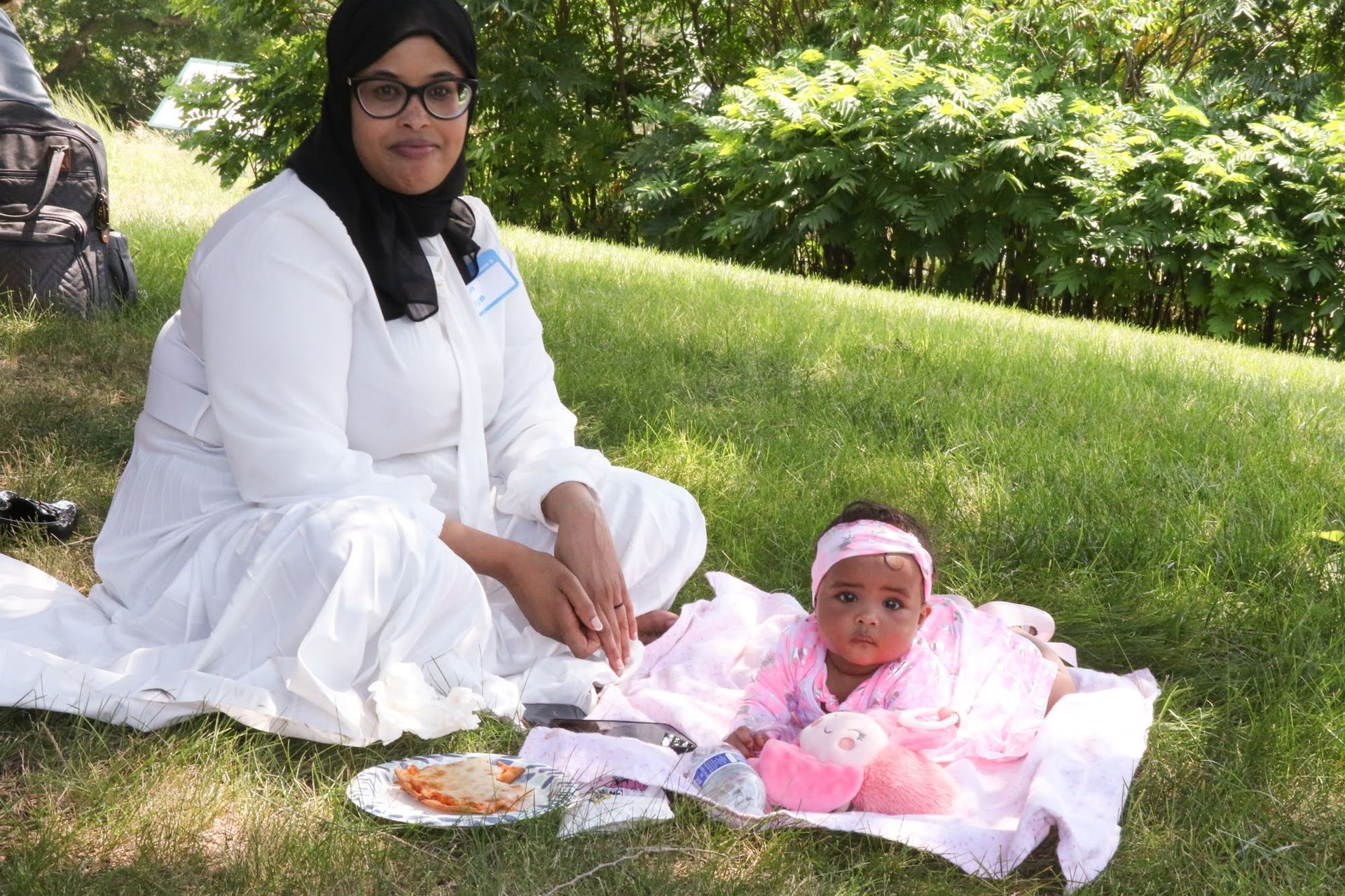 Woman in white attire sits on grass next to baby in pink; outdoor picnic.