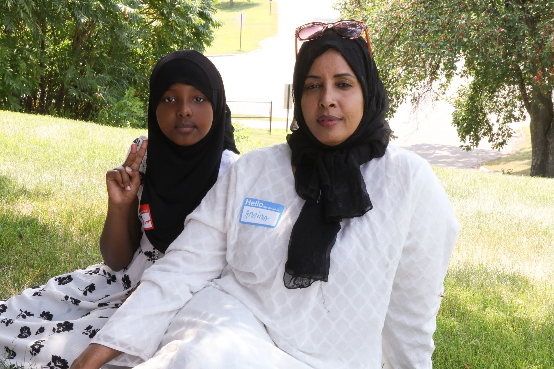 Two people wearing headscarves sit on grassy hill; one points, sunny setting.