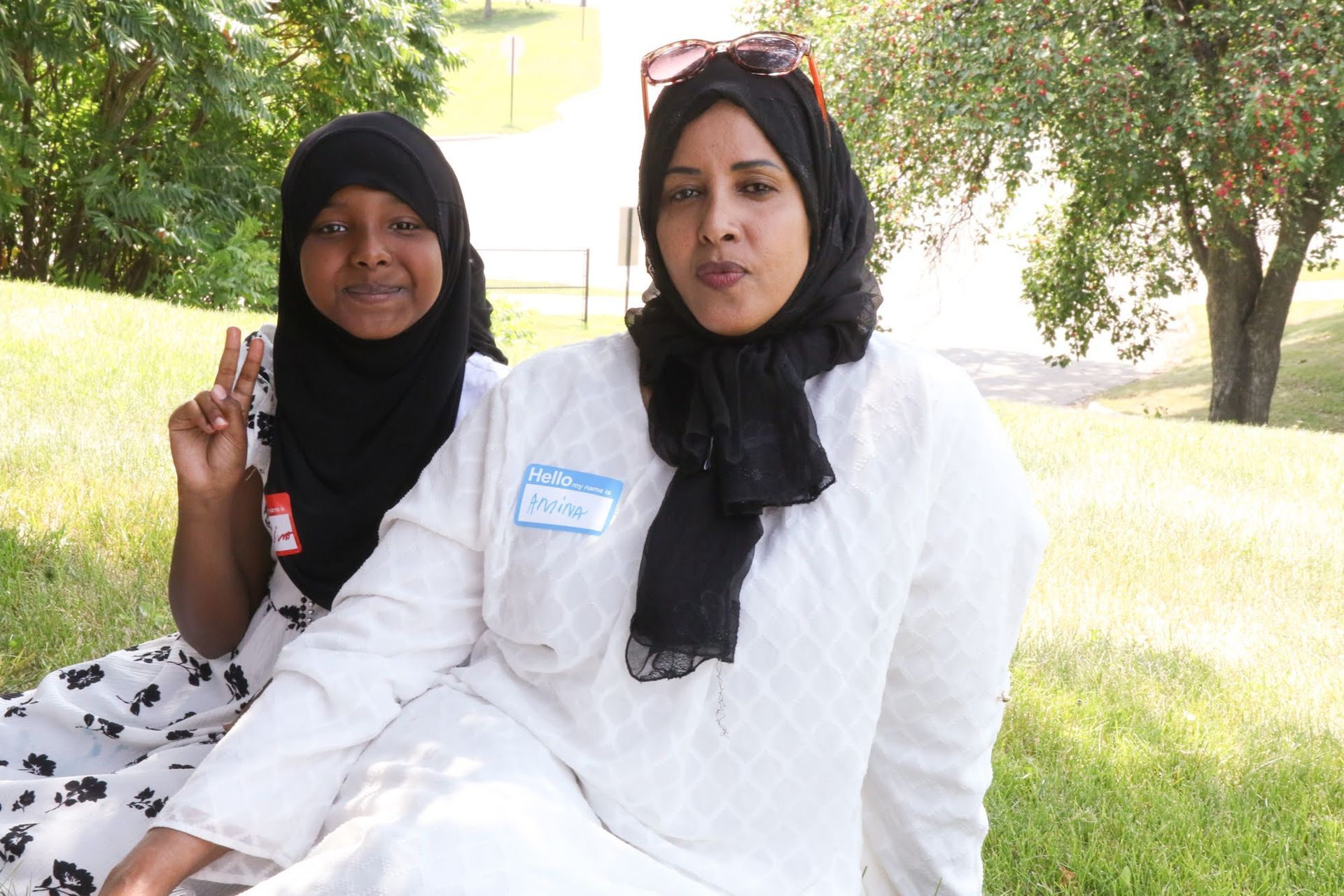 Two women outdoors, one with peace sign. Both wear head coverings. One in white, other in patterned dress.