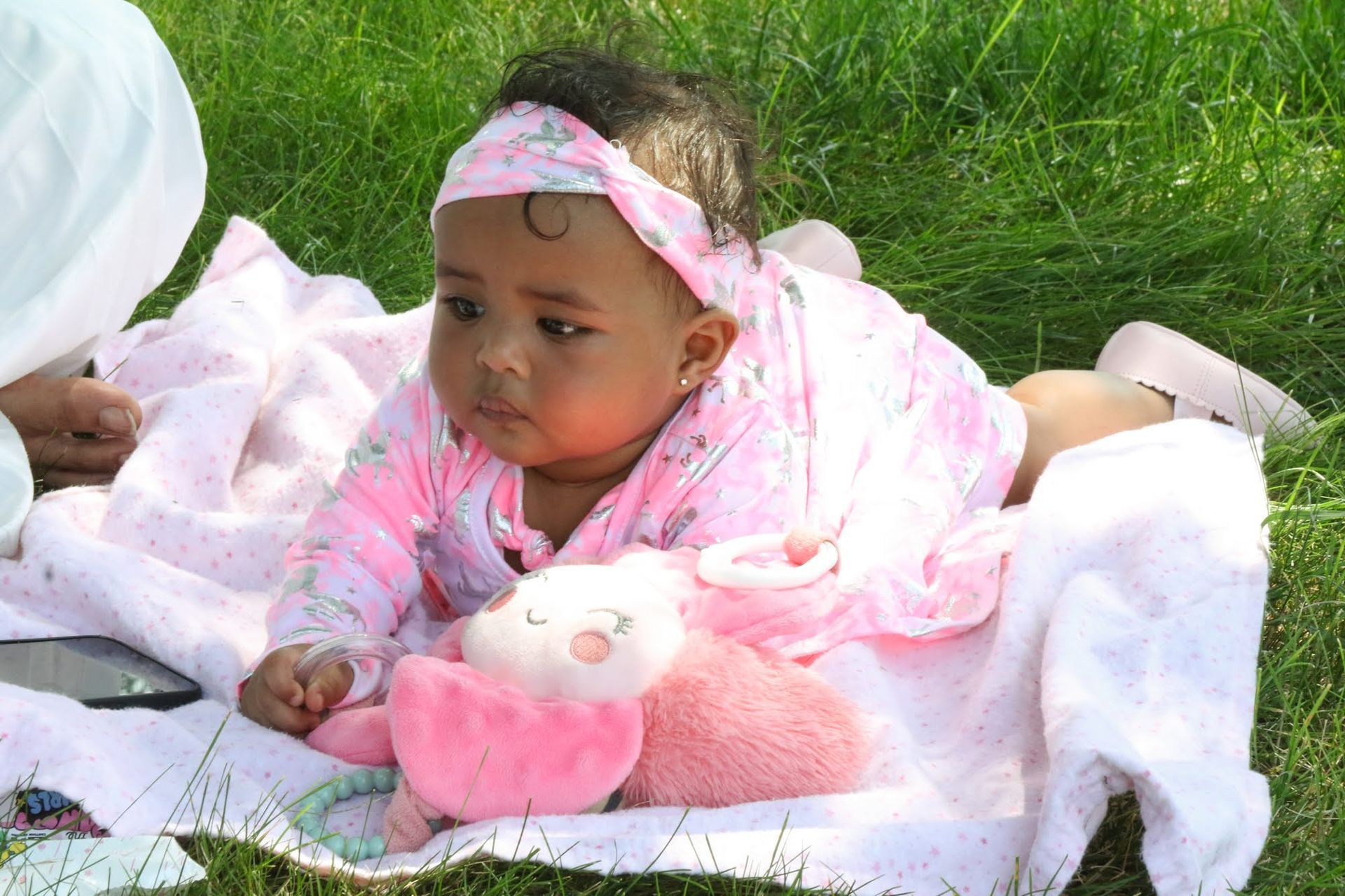 Baby lying on a blanket in grass, wearing a pink outfit and headband, with a stuffed animal.