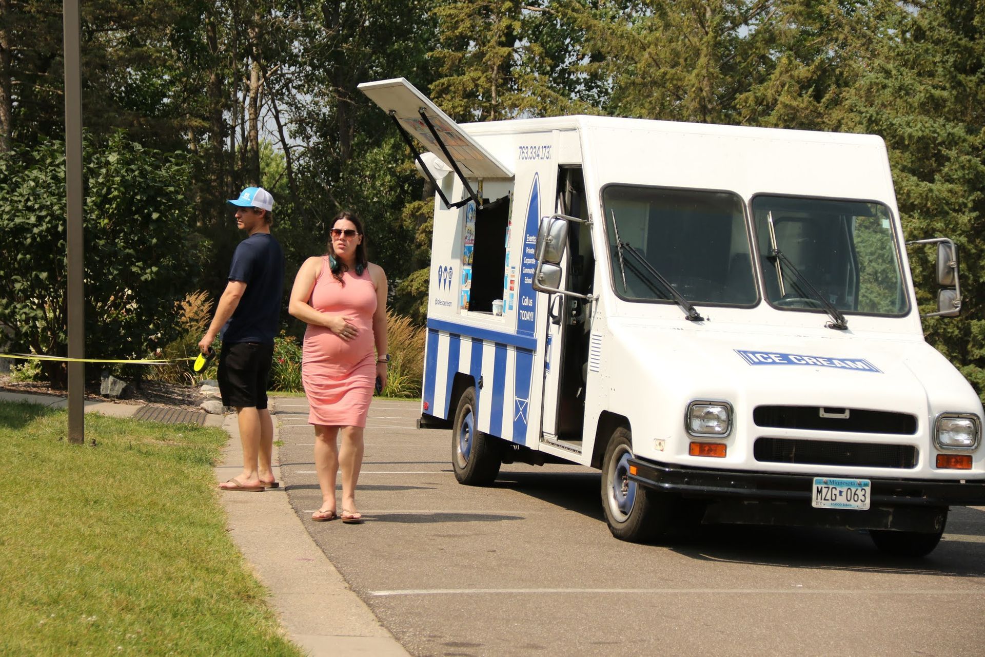 Ice cream truck parked, woman and person wait. Open window shows ice cream selection. Sunny day.