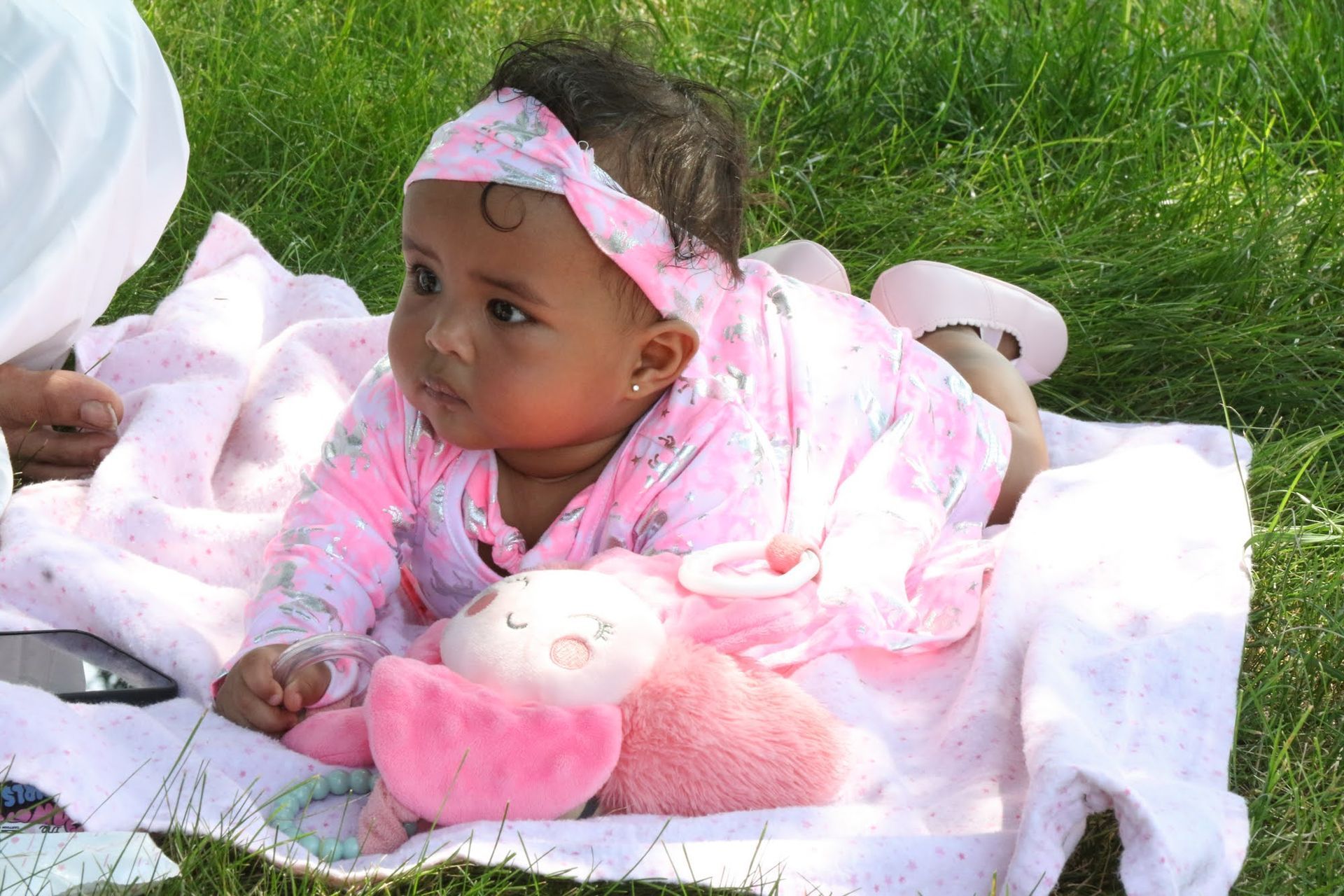 Baby in pink outfit and headband lying on a blanket on the grass, looking off to the side.