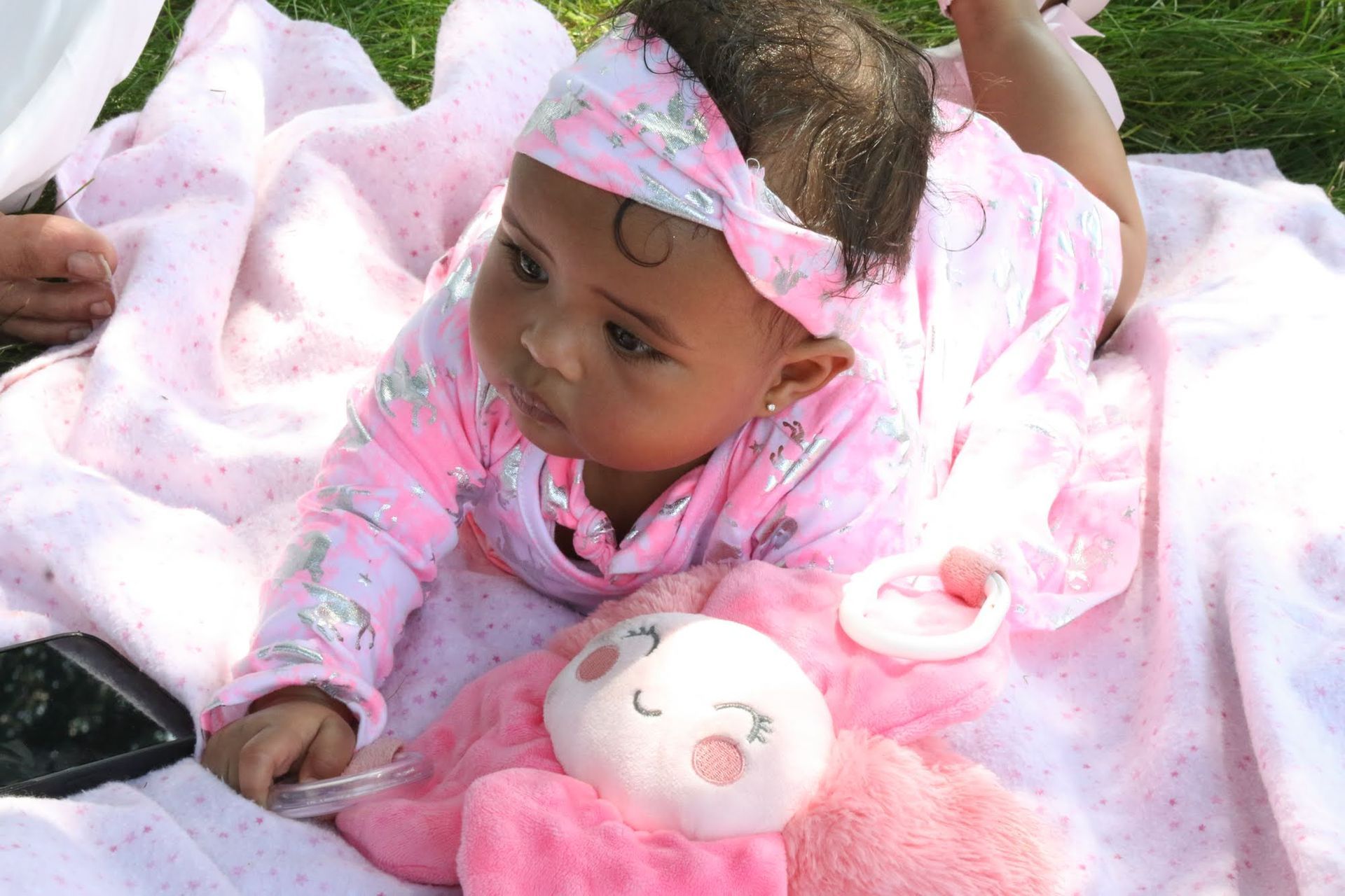 Baby wearing pink outfit, lying on a blanket, looking to the side with a stuffed animal.